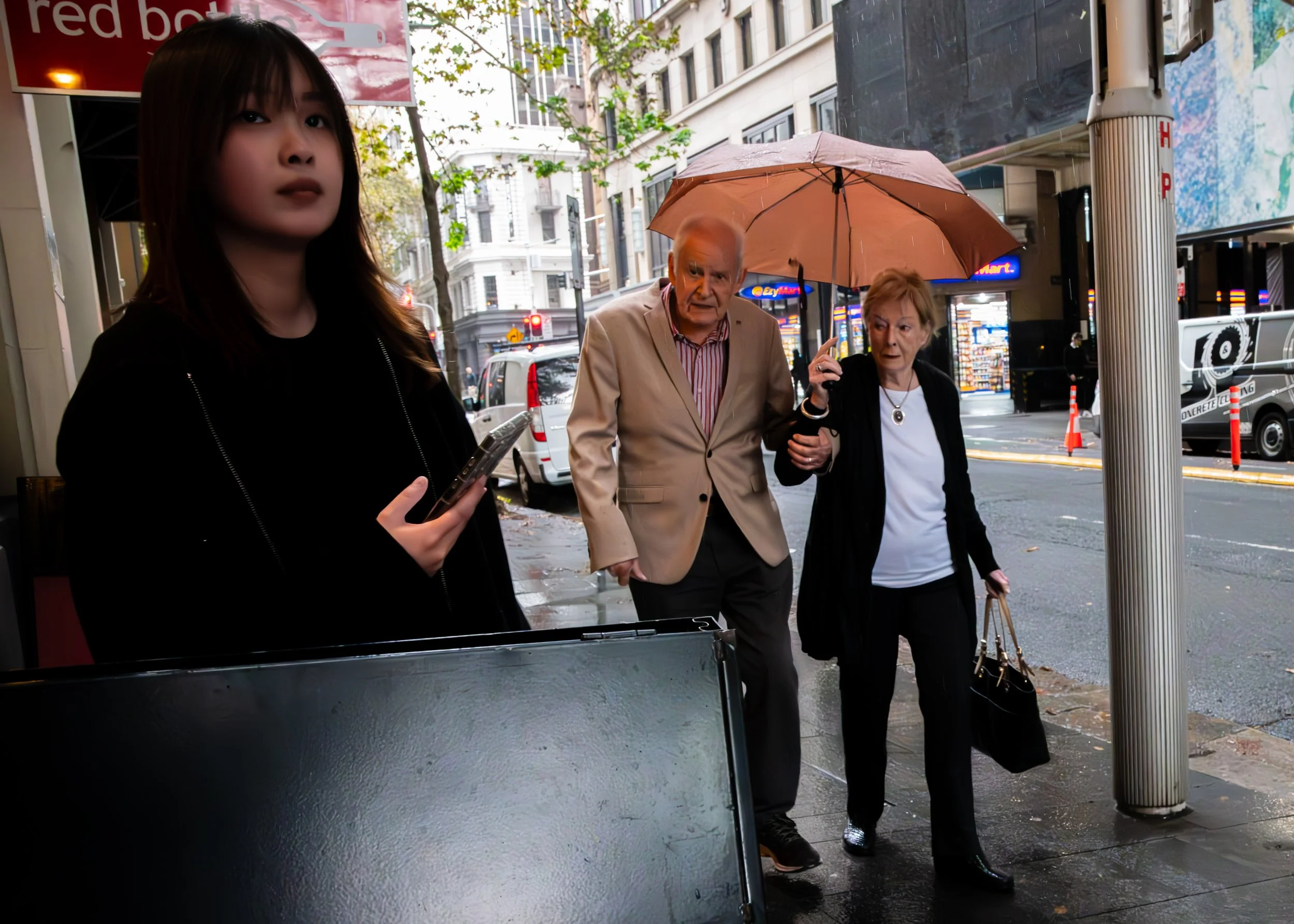 A young woman holding a smartphone stands outside on a city sidewalk next to a black stand. An elderly couple walks past her, with the man carrying an umbrella over both of them. The woman has her arm linked with the man, who looks confused. The street scene includes parked cars, buildings, and bright signs in the background, with some trees and a cloudy sky.