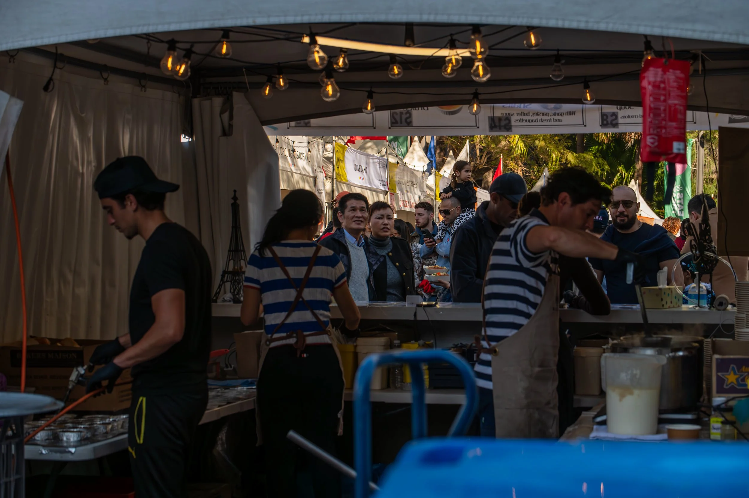 People working at a food stall or outdoor kitchen, with a group of customers on the other side, appearing to enjoy a festival or market setting.