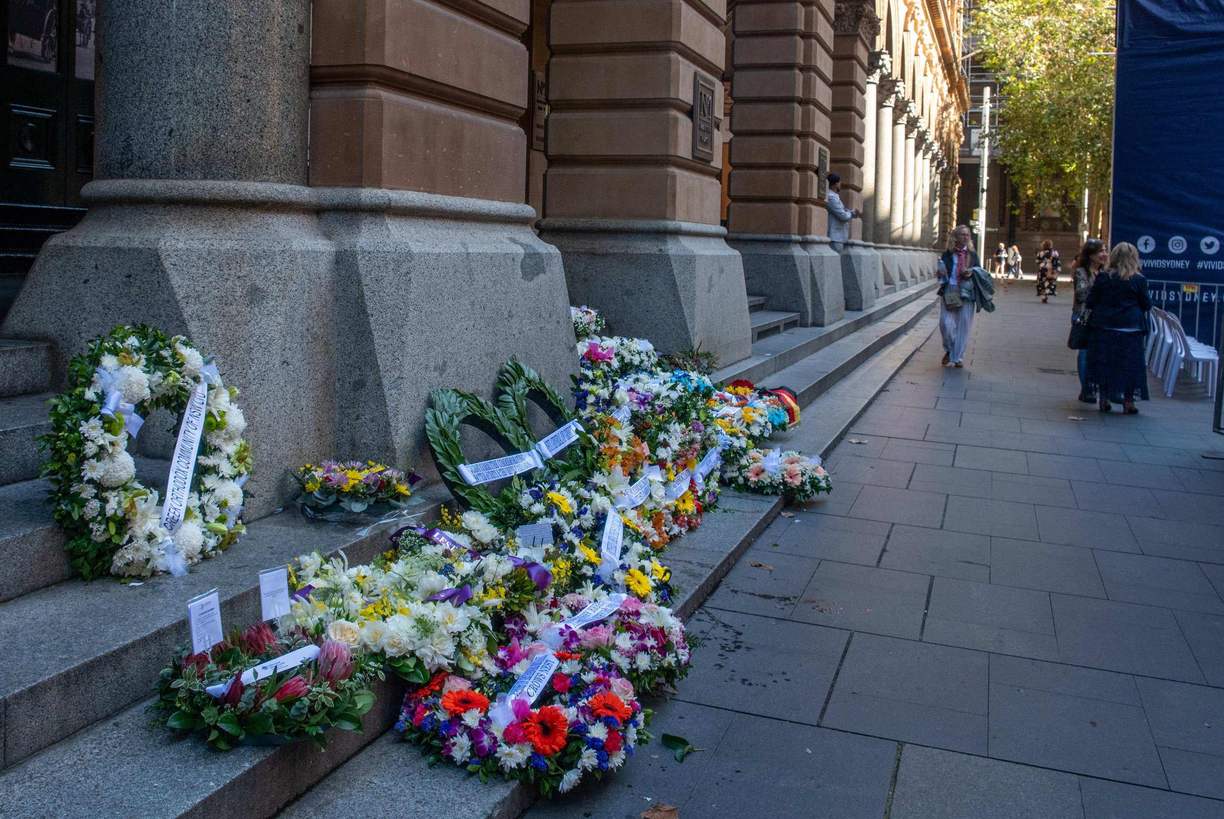 Multiple floral wreaths and bouquets placed on the steps of a large stone building, with a few pedestrians walking nearby.