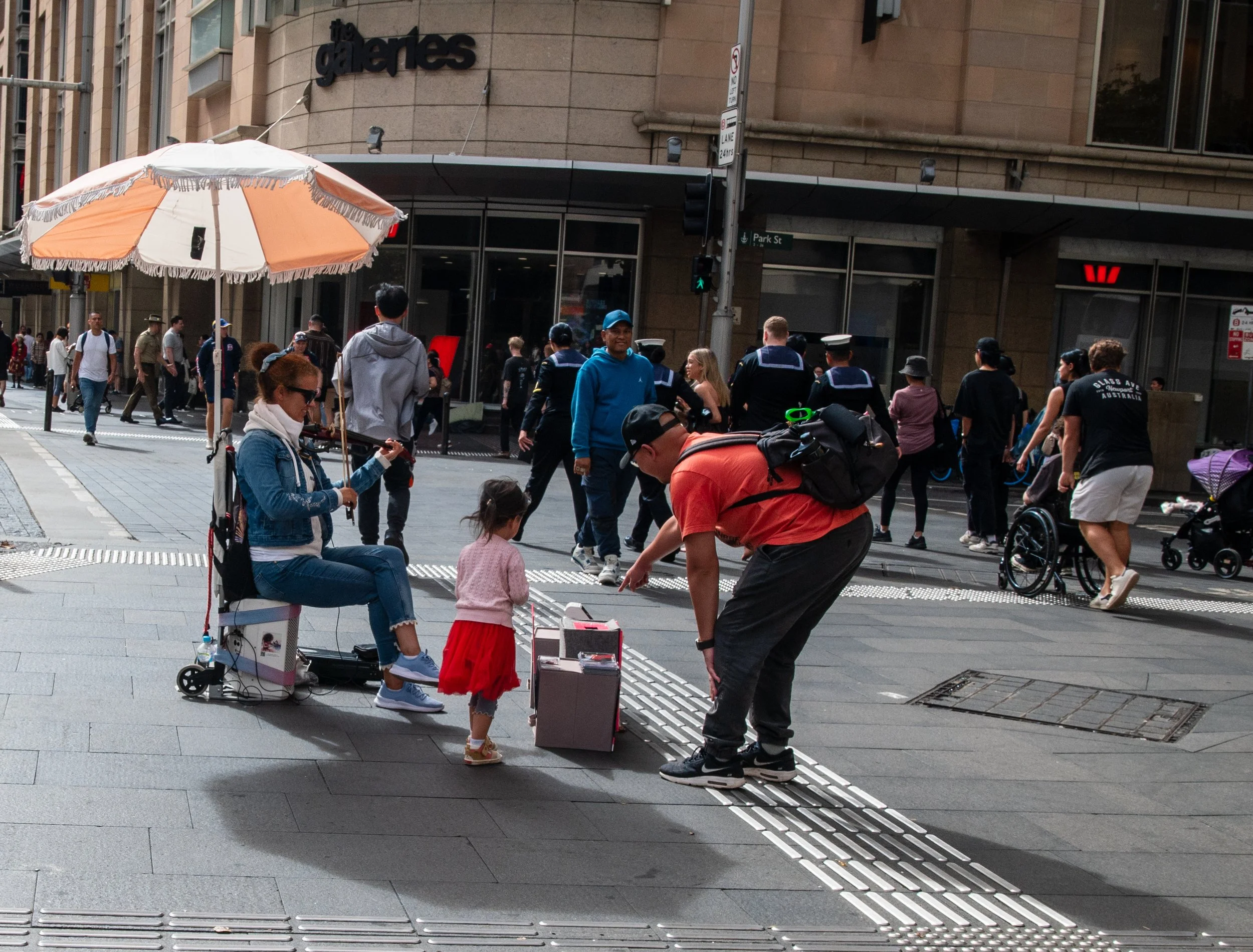 A busy city street with people walking, a seated woman with a small girl, and a man bending down to talk to the girl. The woman appears to be holding an umbrella, and there are storefronts and a building in the background.