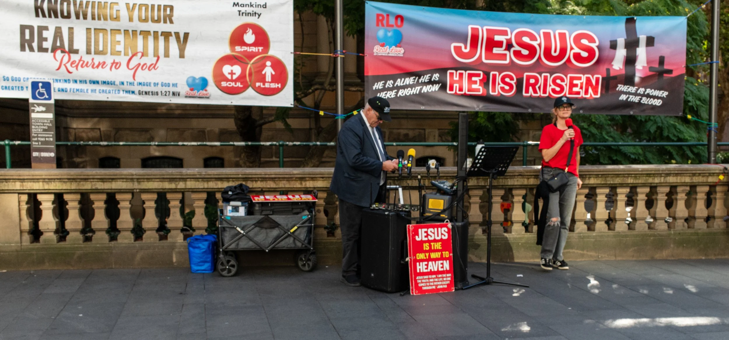 A man with a DJ setup and a woman holding a microphone standing in front of religious banners on a city street.