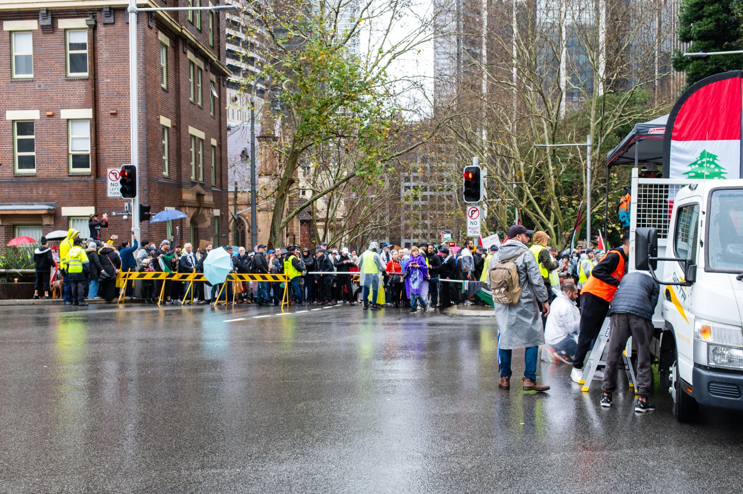 A crowded street scene with a large group of people gathered behind a barricade on a rainy day, some holding umbrellas, near traffic lights and a truck with a Christmas tree banner.