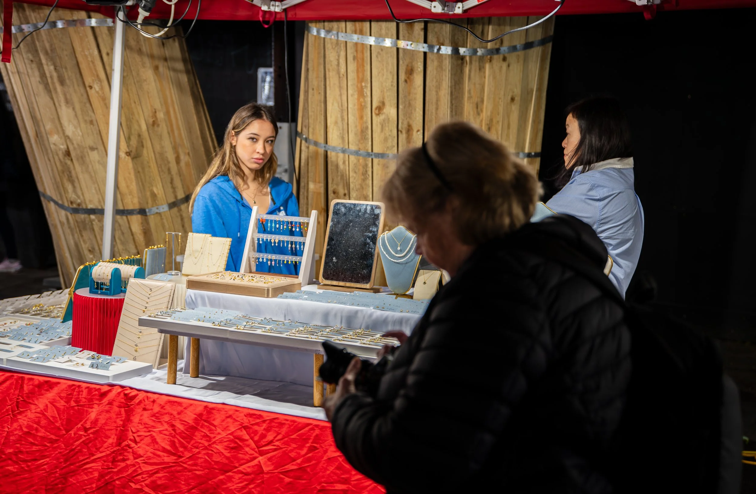 A jewelry booth selling necklaces, earrings, and bracelets at an indoor market, with two women browsing and a woman behind the counter wearing a blue hoodie.