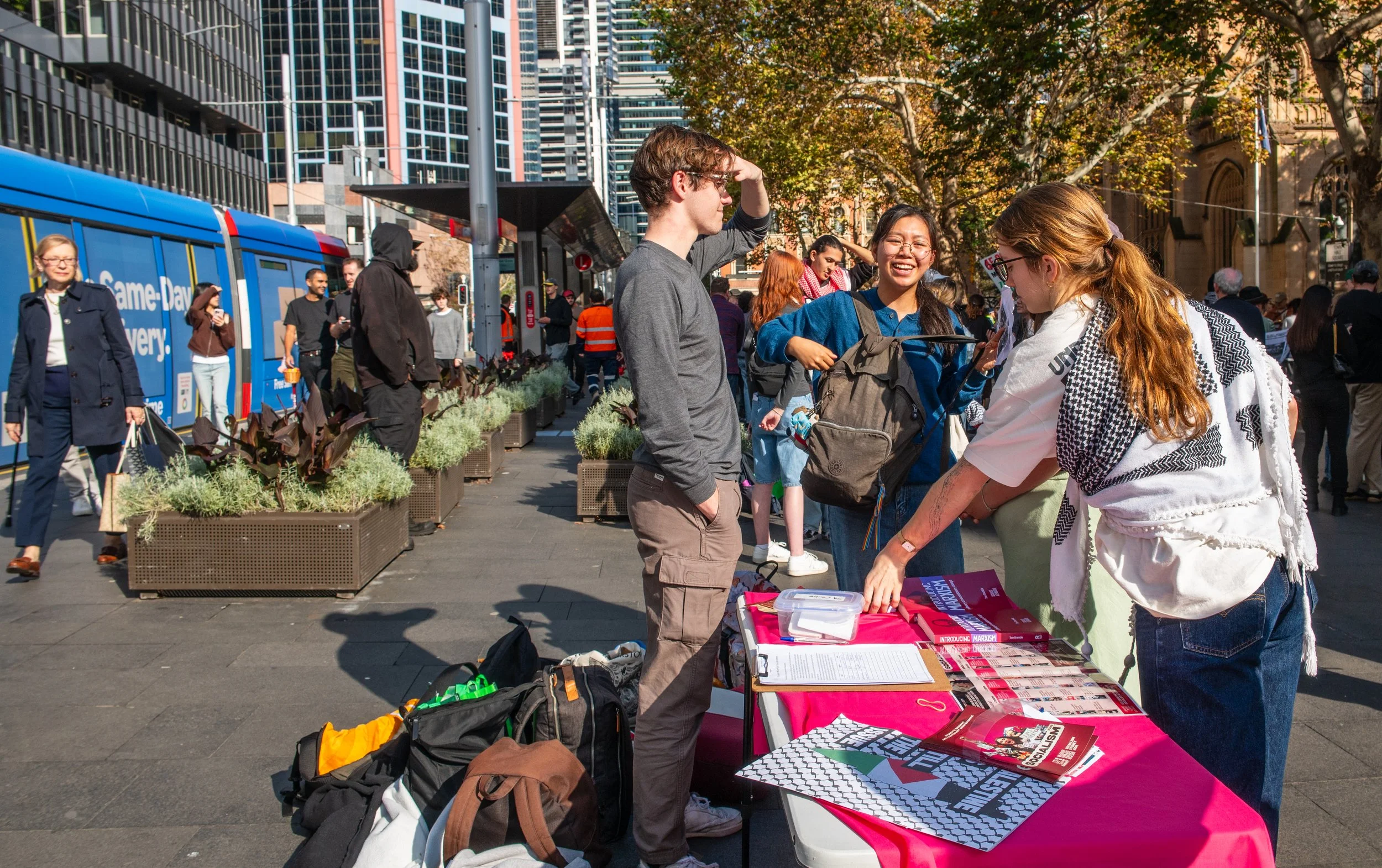 People at an outdoor recruitment or informational table in a city street with trees, tall buildings, and a blue bus in the background, engaging in conversation and browsing informational materials.