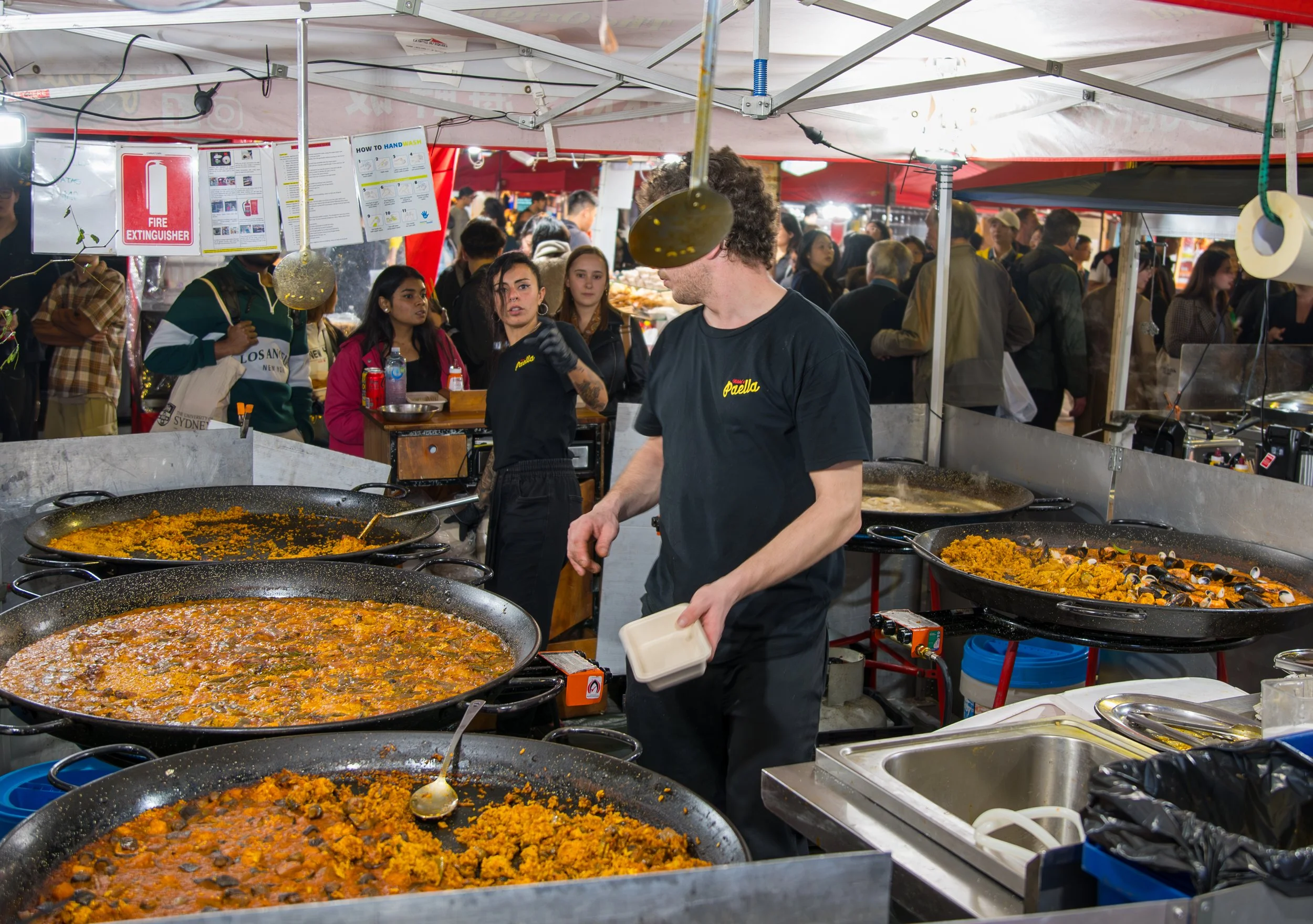 A street food vendor prepares paella in large pans at a busy night market, with a crowd of people shopping in the background.