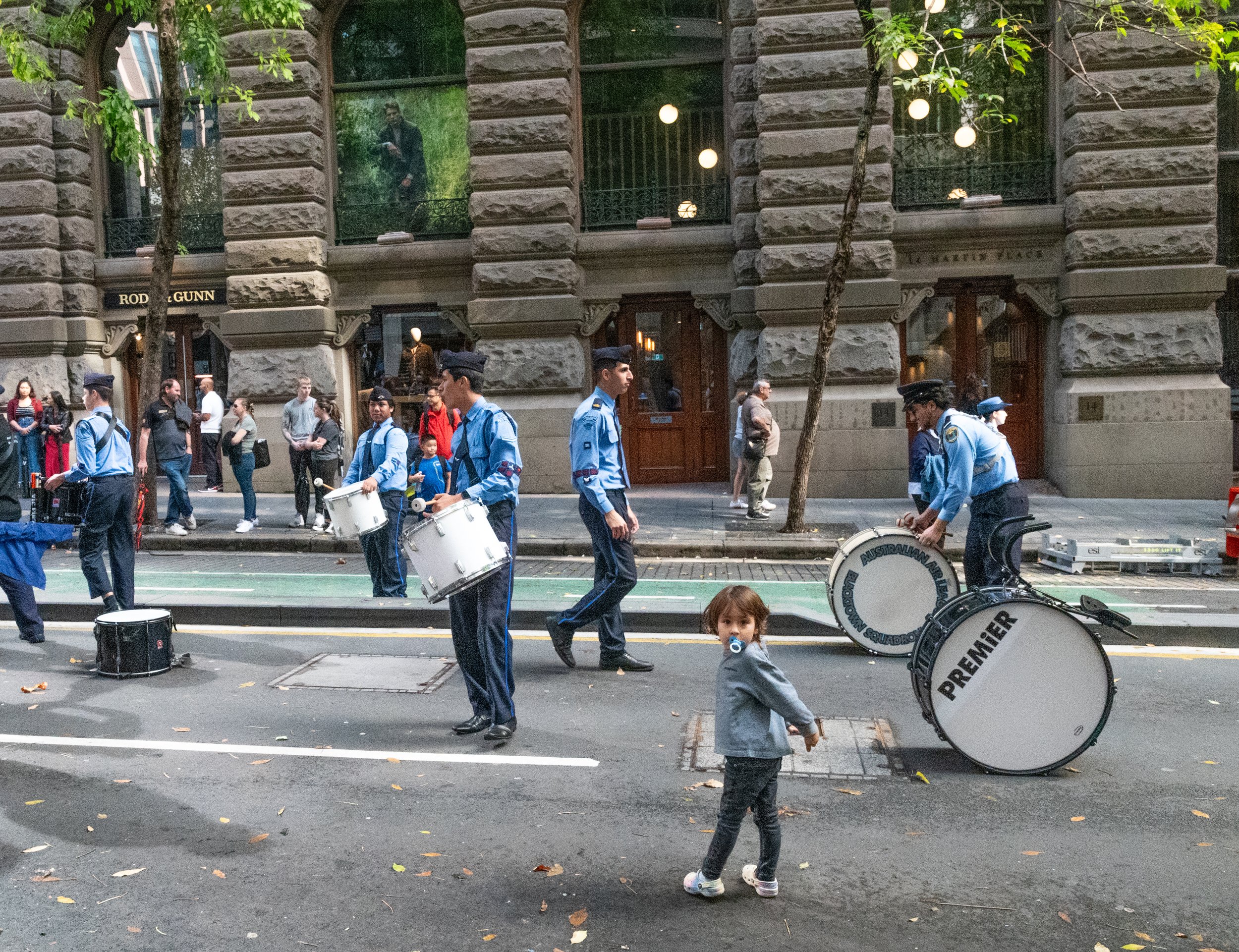 A street scene with a group of uniformed musicians playing drums and other percussion instruments, while pedestrians watch. A young girl with a pacifier stands in the foreground. The background features an ornate stone building and large trees.