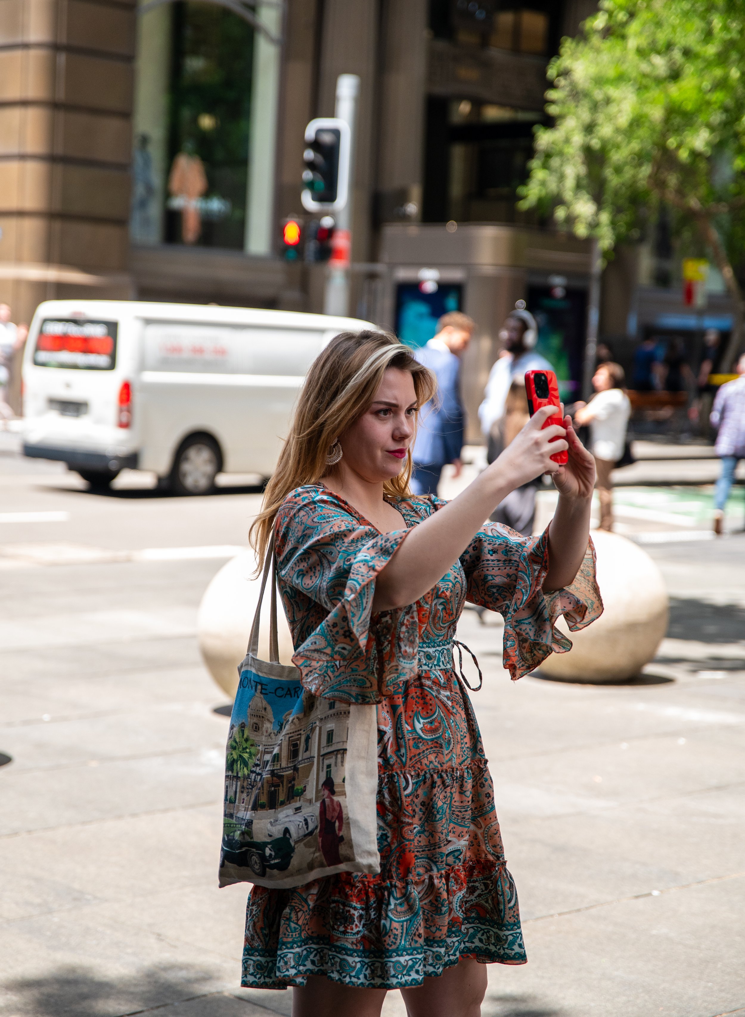 A woman is standing on a city sidewalk taking a selfie or photo with a red smartphone. She has long blonde hair, wears earrings, a colorful patterned dress, and carries a tote bag with a vintage car and city scene printed on it. Behind her, there are pedestrians, a parked white van, and tall buildings.