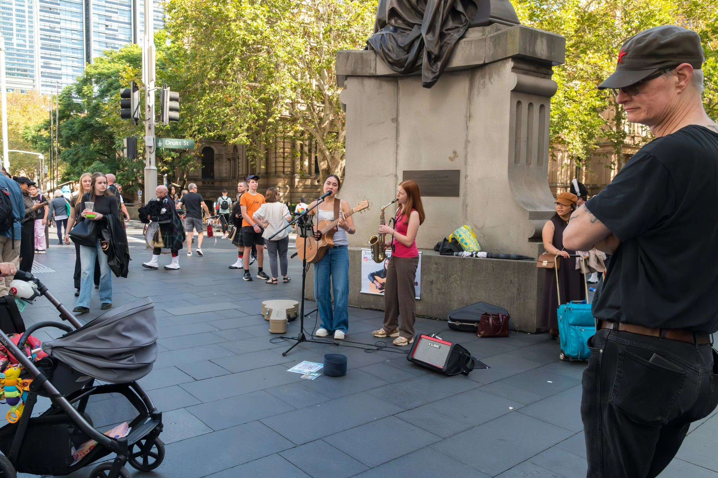 Two women perform music with guitar and saxophone on a street corner while a crowd watches. A man with sunglasses and a cap stands nearby. The scene takes place outdoors in an urban setting with trees and city buildings in the background.
