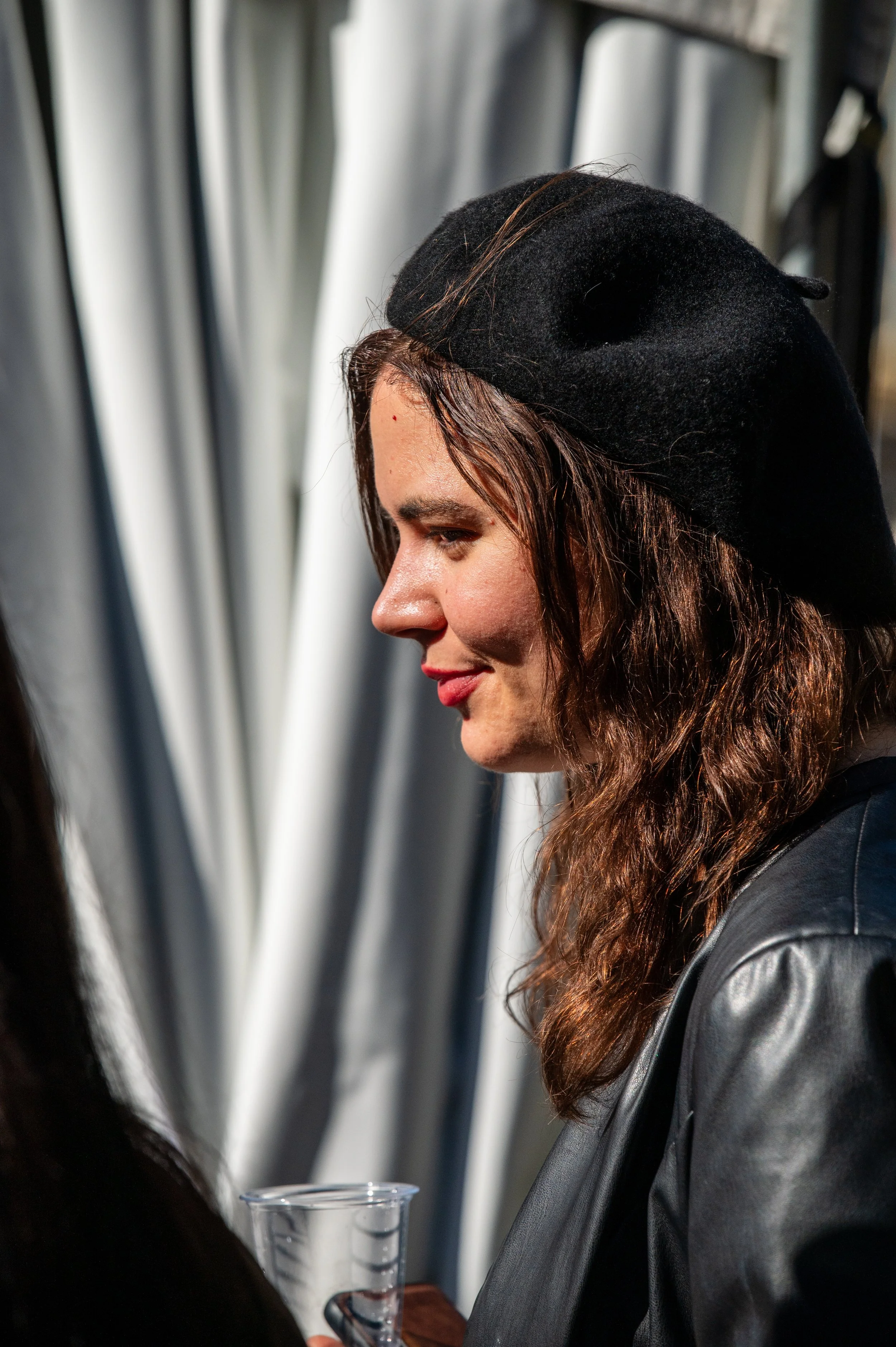 Side profile of a woman with wavy brown hair wearing a black hat and black leather jacket, holding a clear plastic cup.