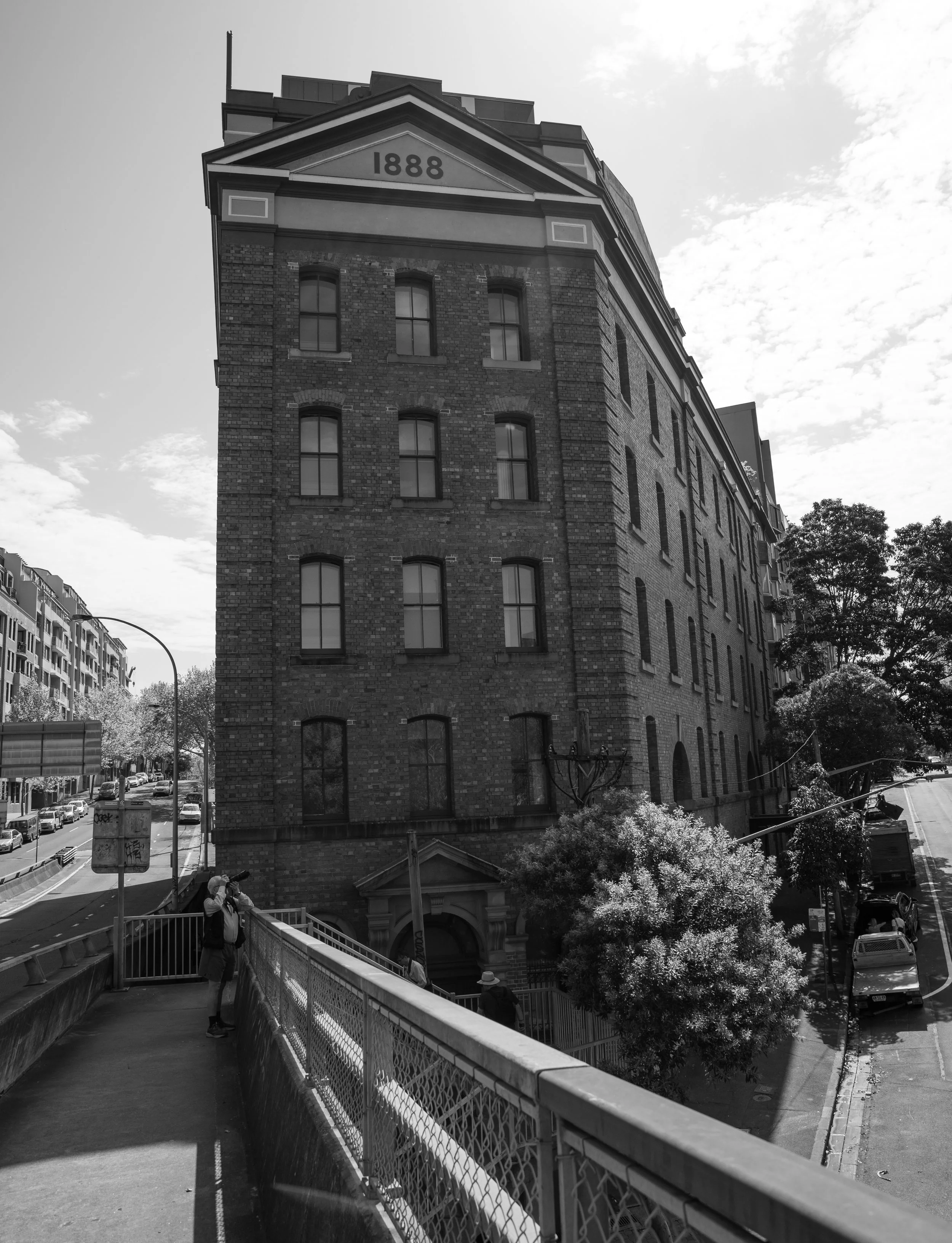 Black and white photo of an old brick building with the year 1888 inscribed on the facade, multiple windows, and a tree in front. A person is taking a photo near a railing, with a city street in the background.