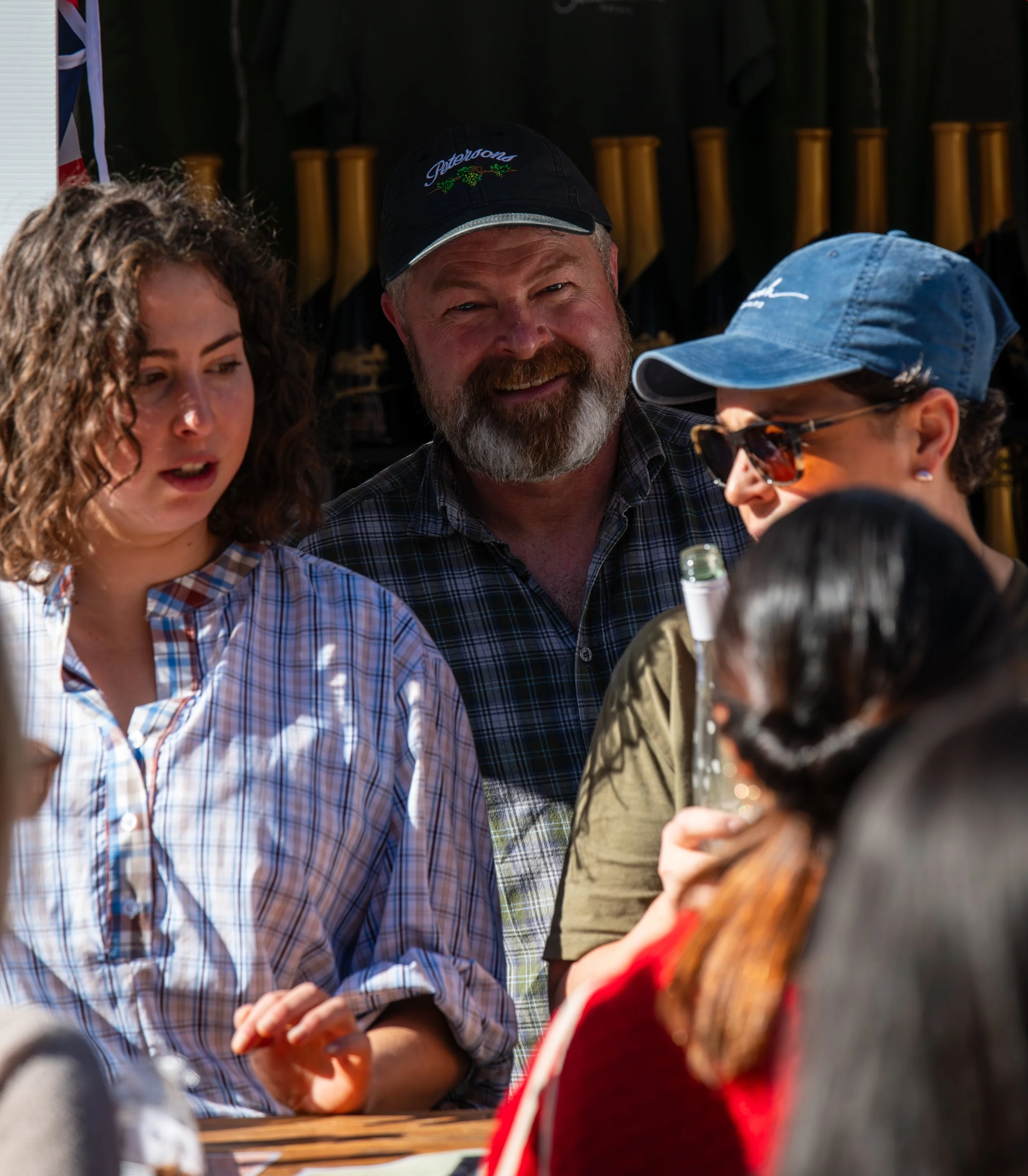 Group of people gathered outdoors, engaged in conversation, with shelves of bottles in the background.