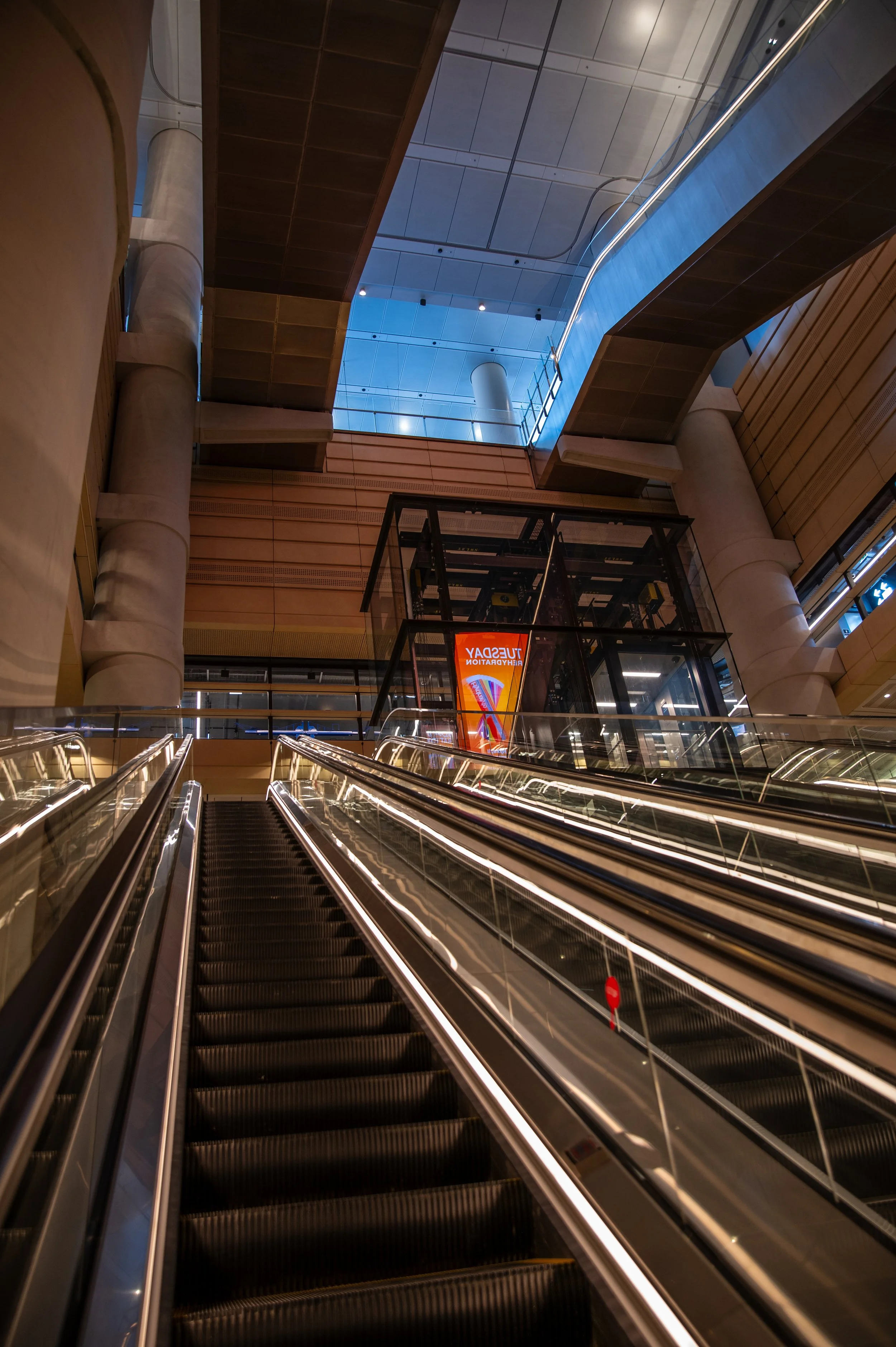 Interior view of a modern building with escalators leading to upper levels, large wooden and concrete structural elements, and a glass elevator shaft with a colorful poster inside.
