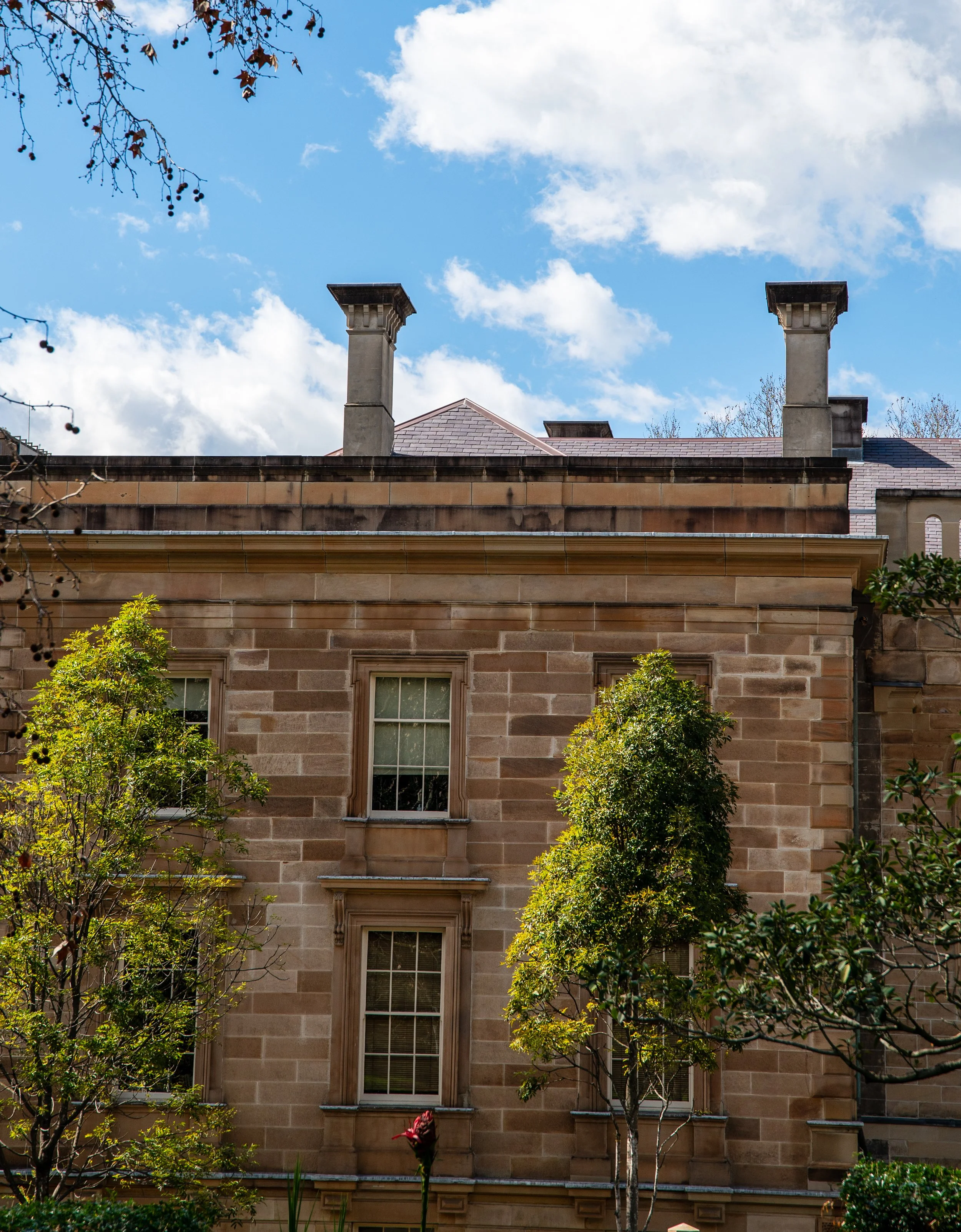 A brick building with two trees in front and two chimneys on the roof, under a partly cloudy blue sky.