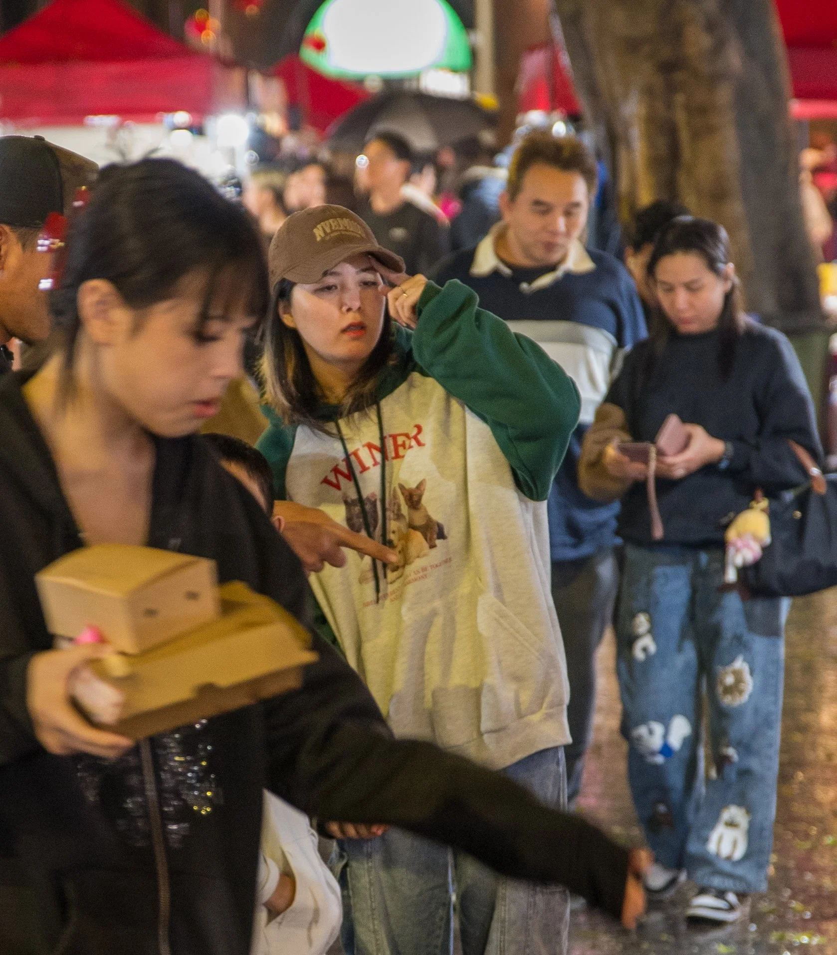 A group of people standing in line outdoors at night, with some looking at their phones, a woman in the foreground holding food boxes, and others waiting behind her.