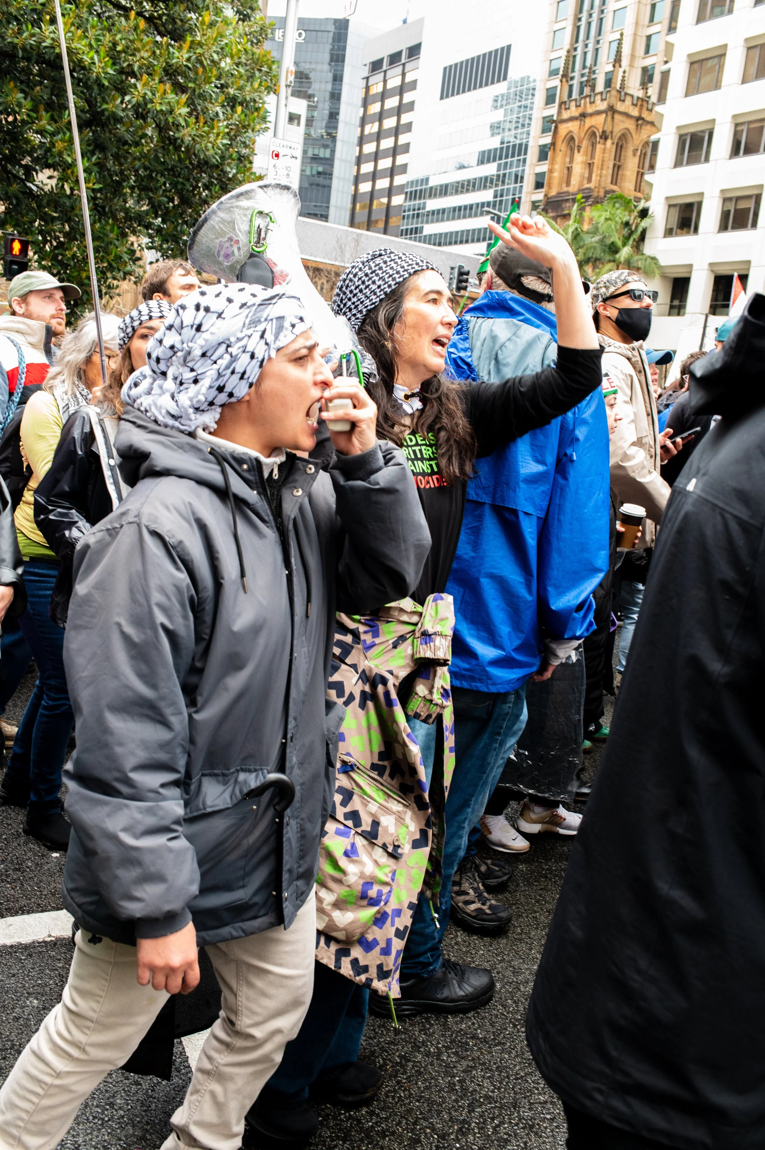 People protesting on a city street, some wearing masks and rain jackets, with one woman passionately speaking and raising her hand.