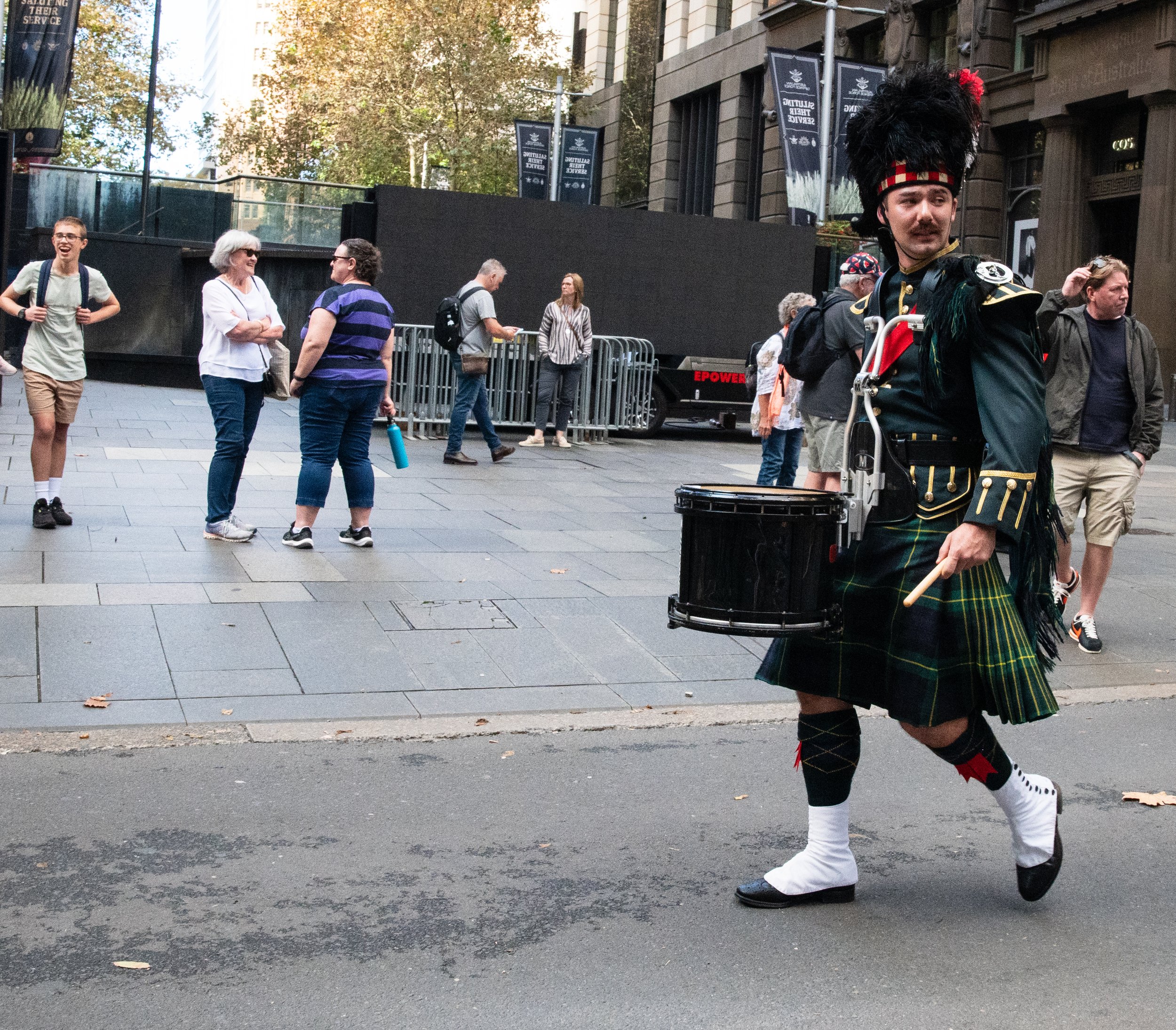 A man dressed as a Scottish bagpiper walking on a city street lined with buildings, with a group of casually dressed people talking and walking in the background.
