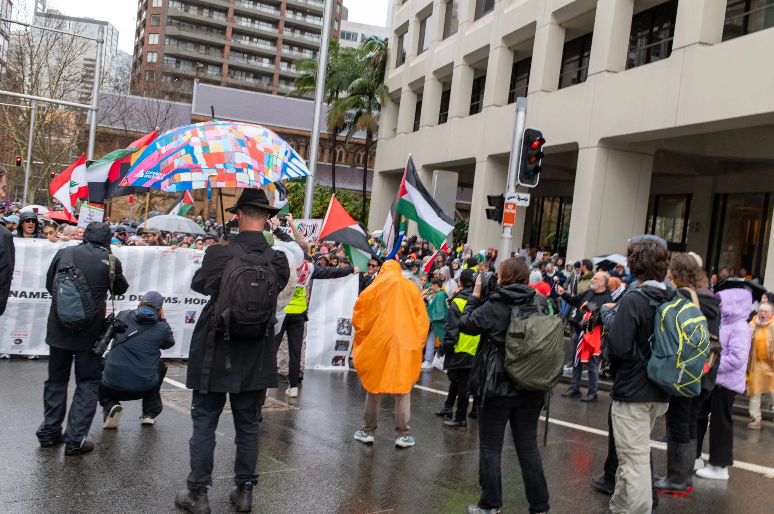 A group of people participating in a protest on a rainy day, holding flags and banners, with some wearing raincoats and umbrellas.