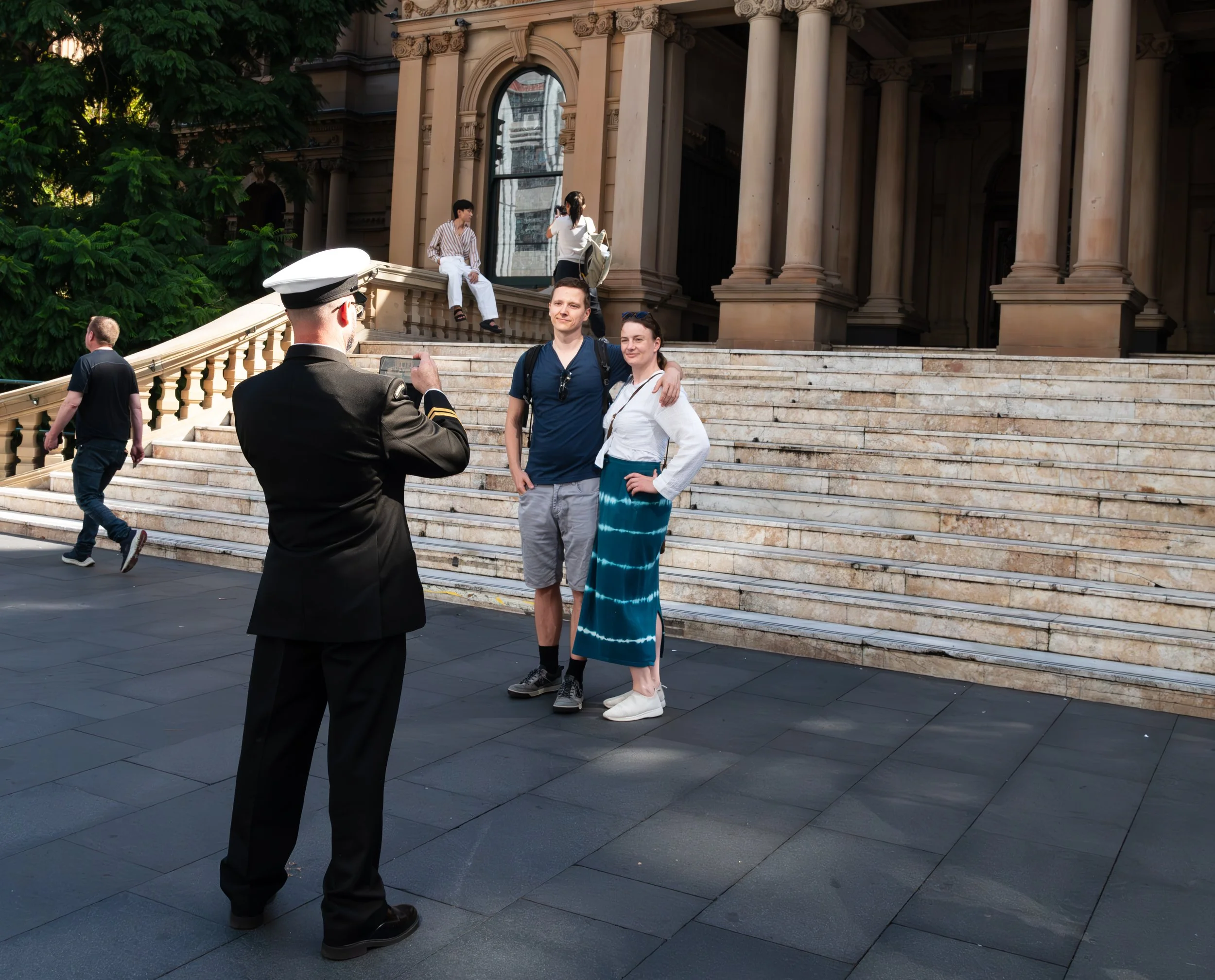 A man in a military uniform taking a photo of a smiling couple on the steps of a historic building, with other pedestrians and photographers in the background.