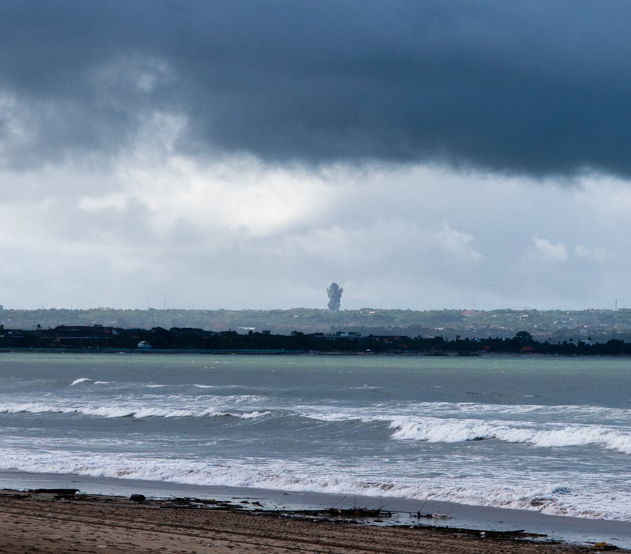 A beach scene with waves crashing onto the shore, overcast sky with dark clouds, and a distant city skyline with tall buildings visible in the background.