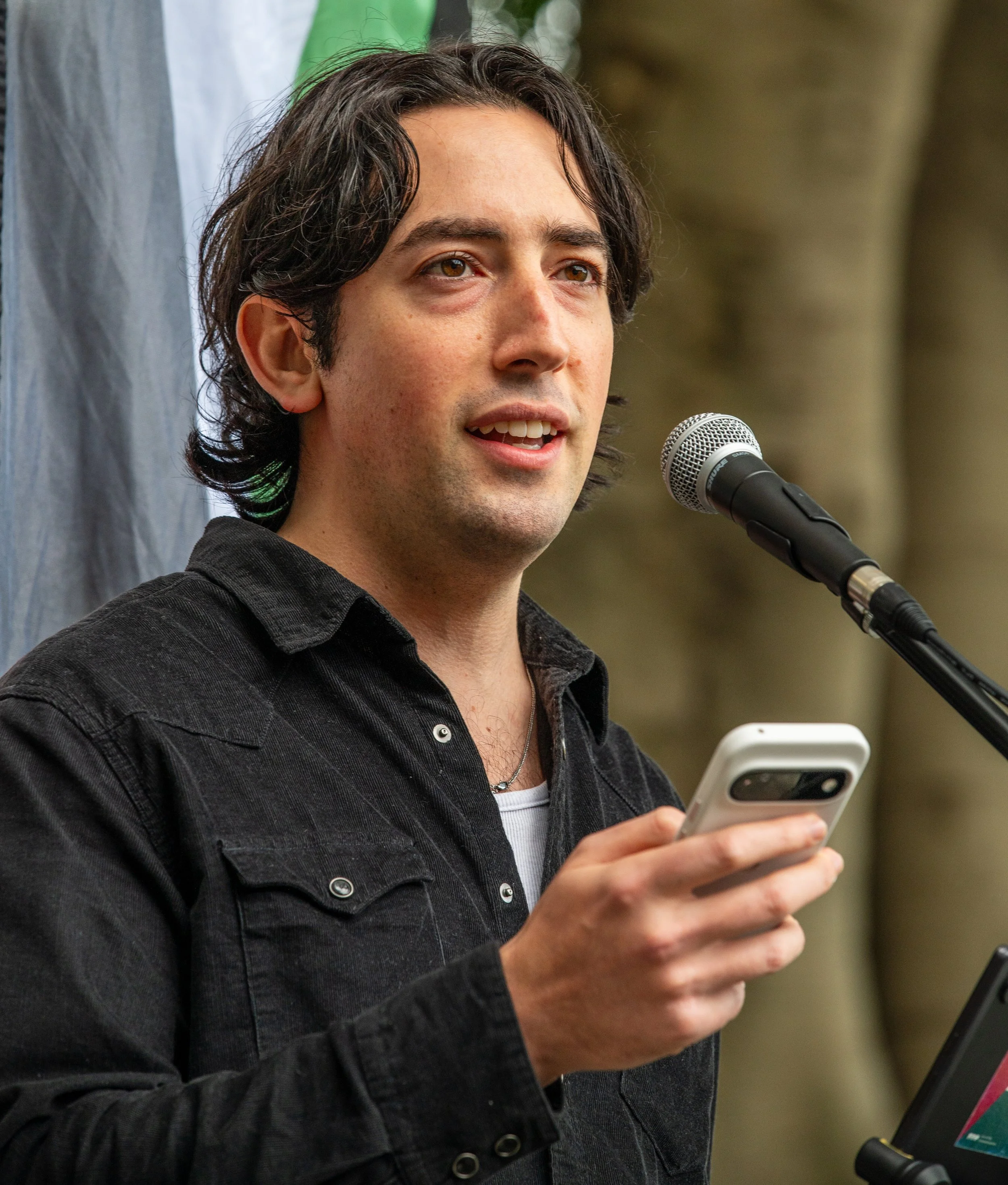 Young man speaking into a microphone while holding a smartphone at an outdoor event.