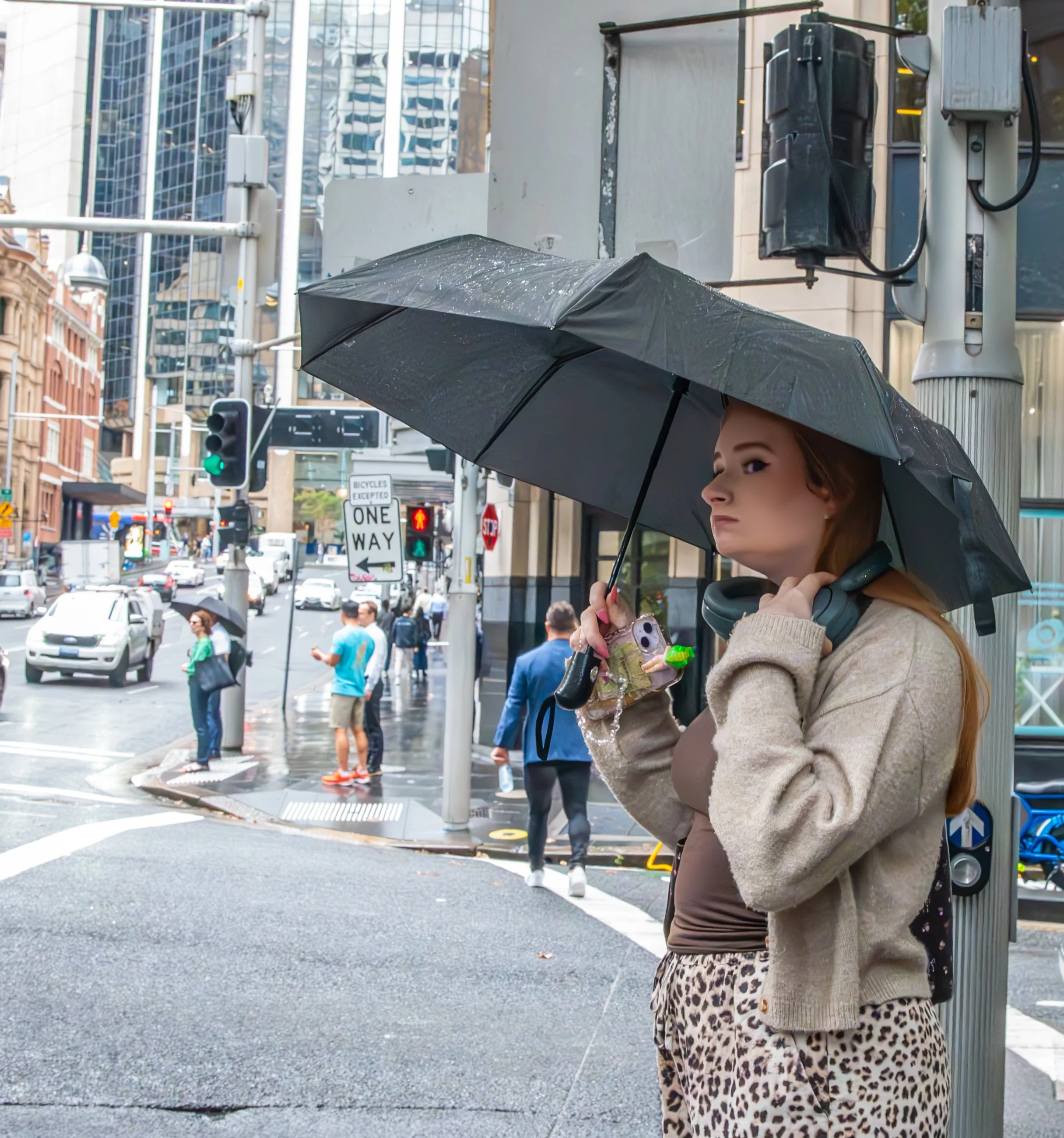A woman with red hair standing at a city crosswalk holding a black umbrella in the rain, wearing a beige jacket, leopard print pants, and headphones around her neck, with city buildings and other pedestrians in the background.