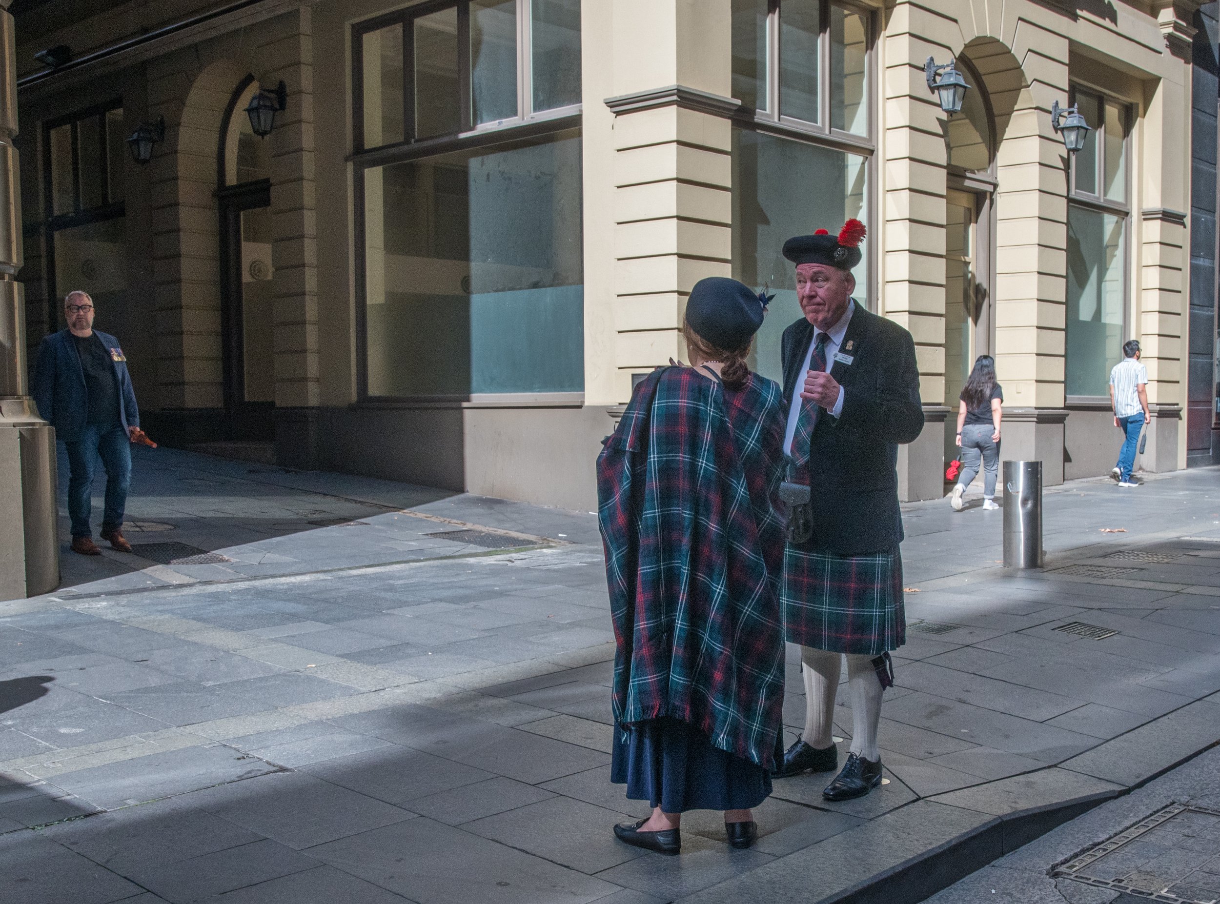 Two individuals dressed in traditional Scottish attire, including kilts and tartan shawls, are talking on a city street, with one man wearing a hat with a red feather. A man in modern casual clothing is walking in the background.