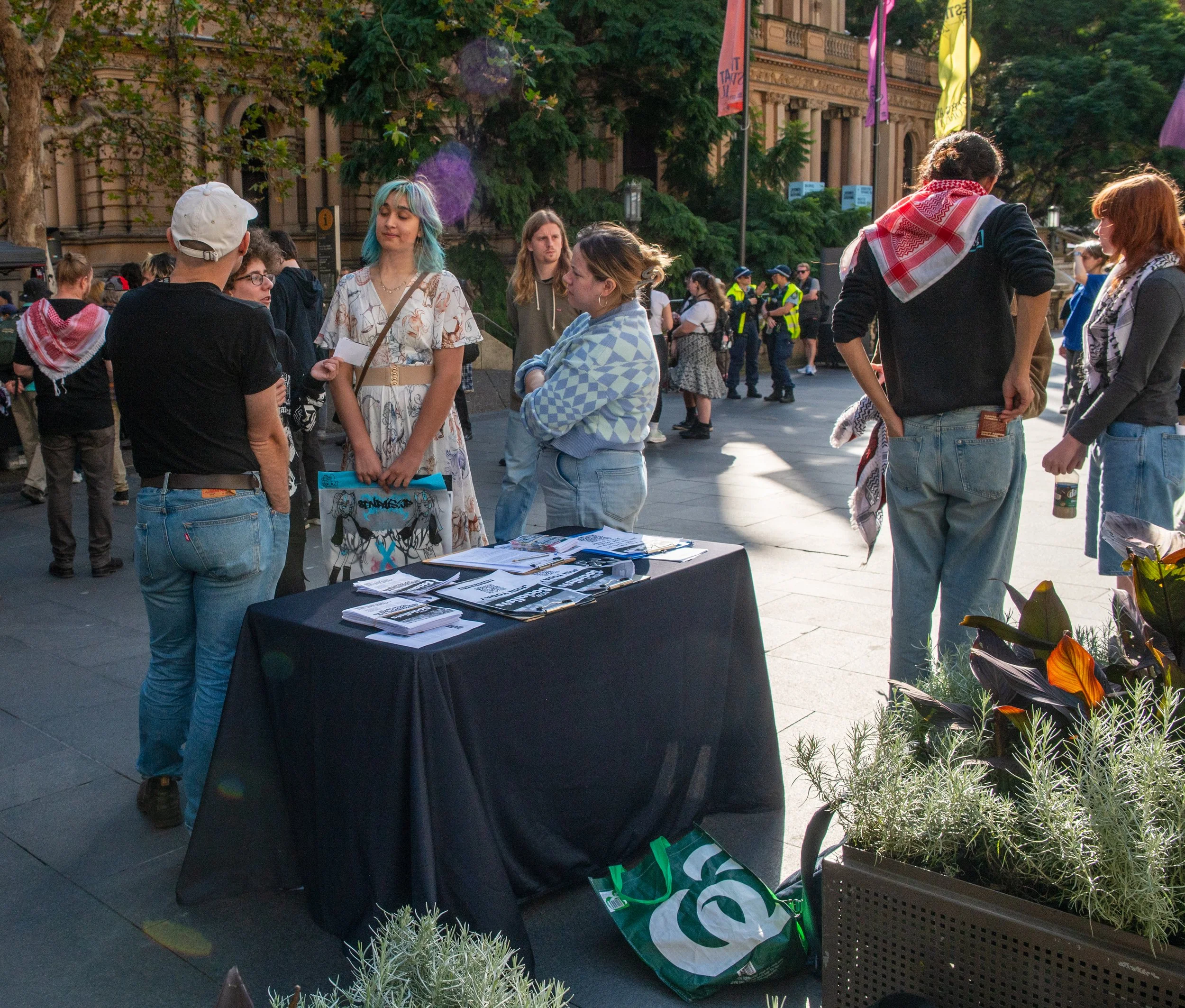Group of people gathered around a table with pamphlets and flyers outdoors, with police officers and a historic building in the background.