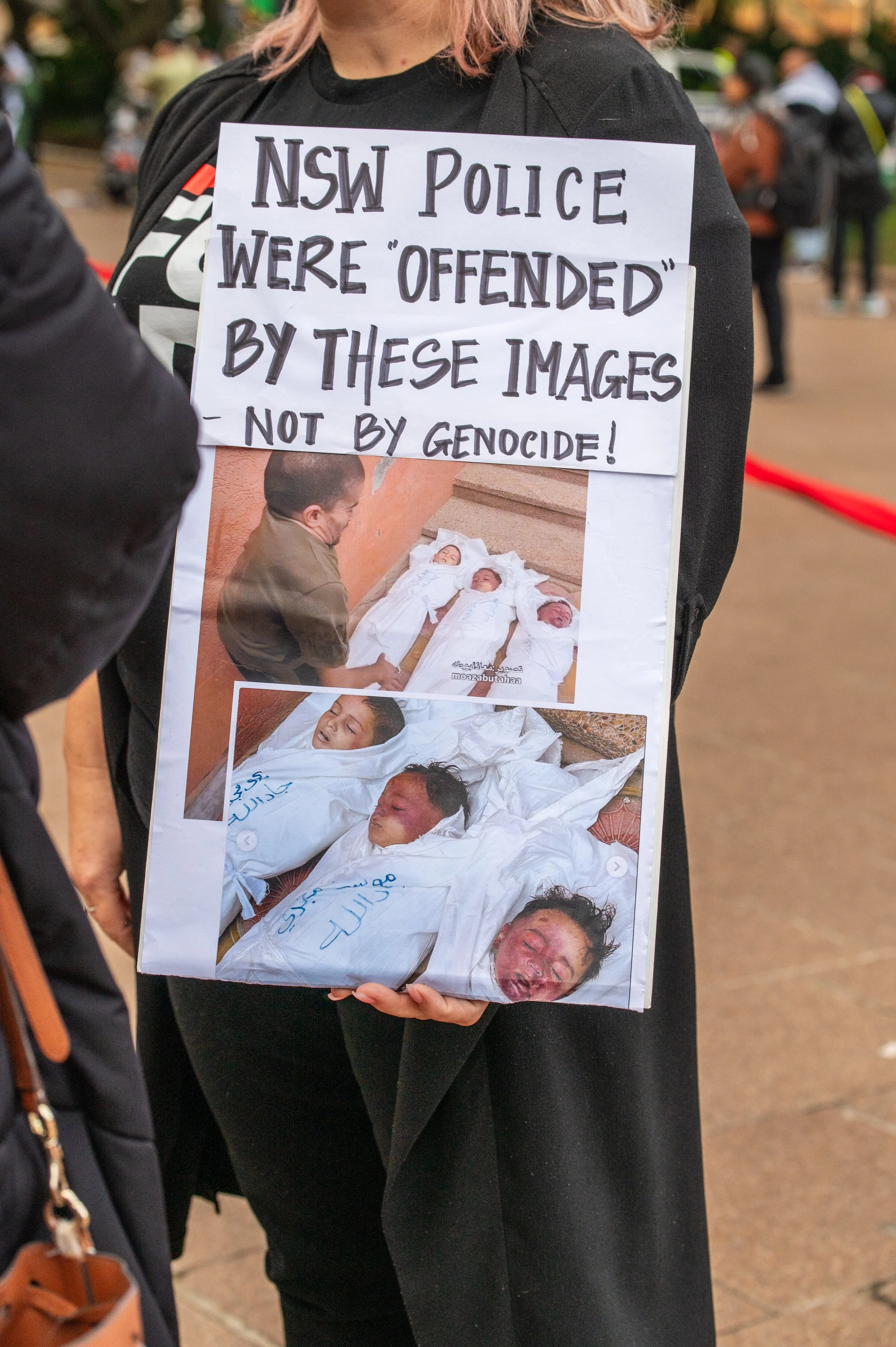 Person holding a protest sign with photos of victims of violence and a message criticizing the NSW Police for being offended by images, not by genocide.