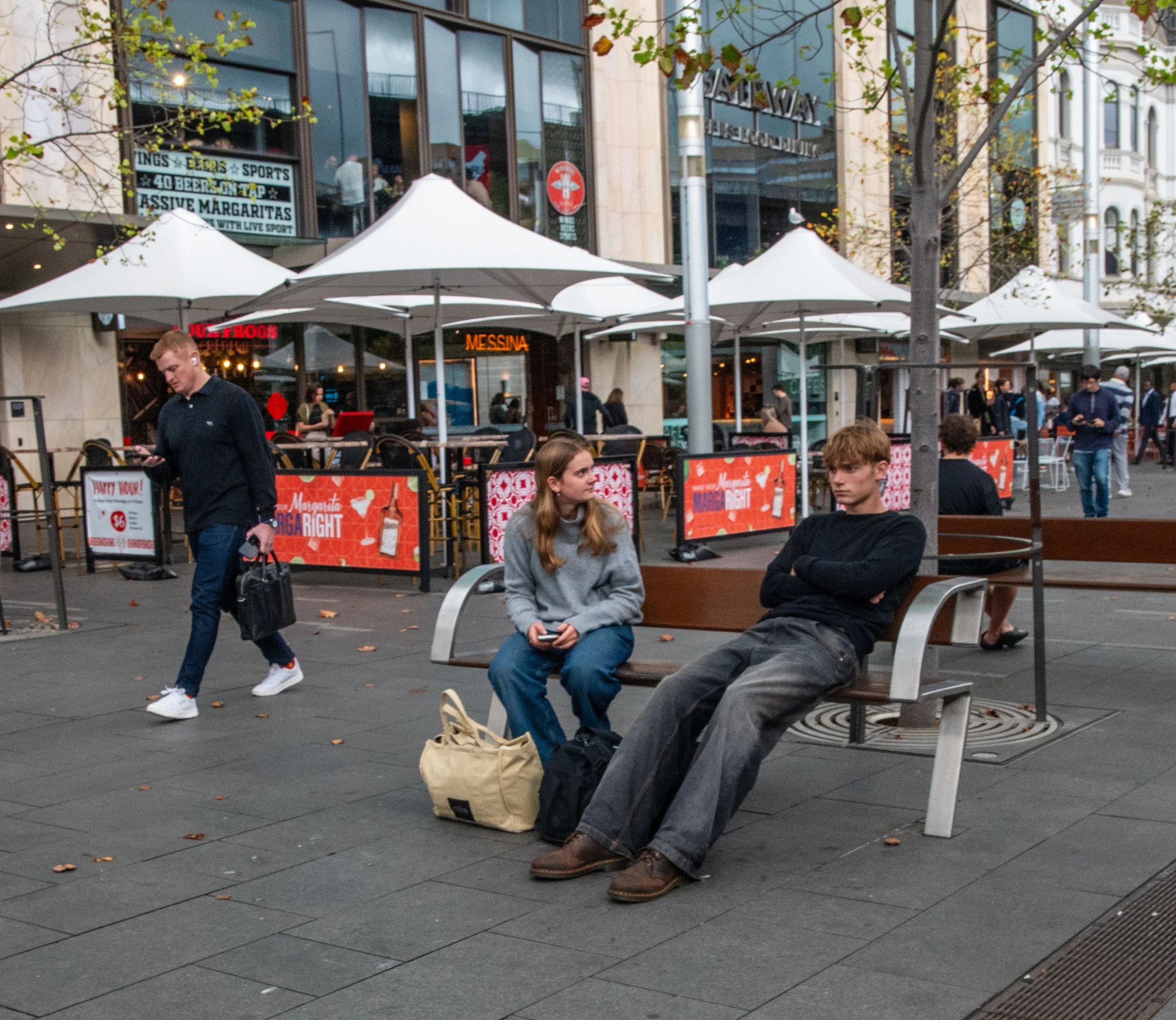 Two young people sitting on a bench on a city sidewalk, engaged in conversation, with a shopping bag and backpack nearby. Behind them, a busy outdoor dining area with white umbrellas and people walking, in front of a modern building with large glass windows.