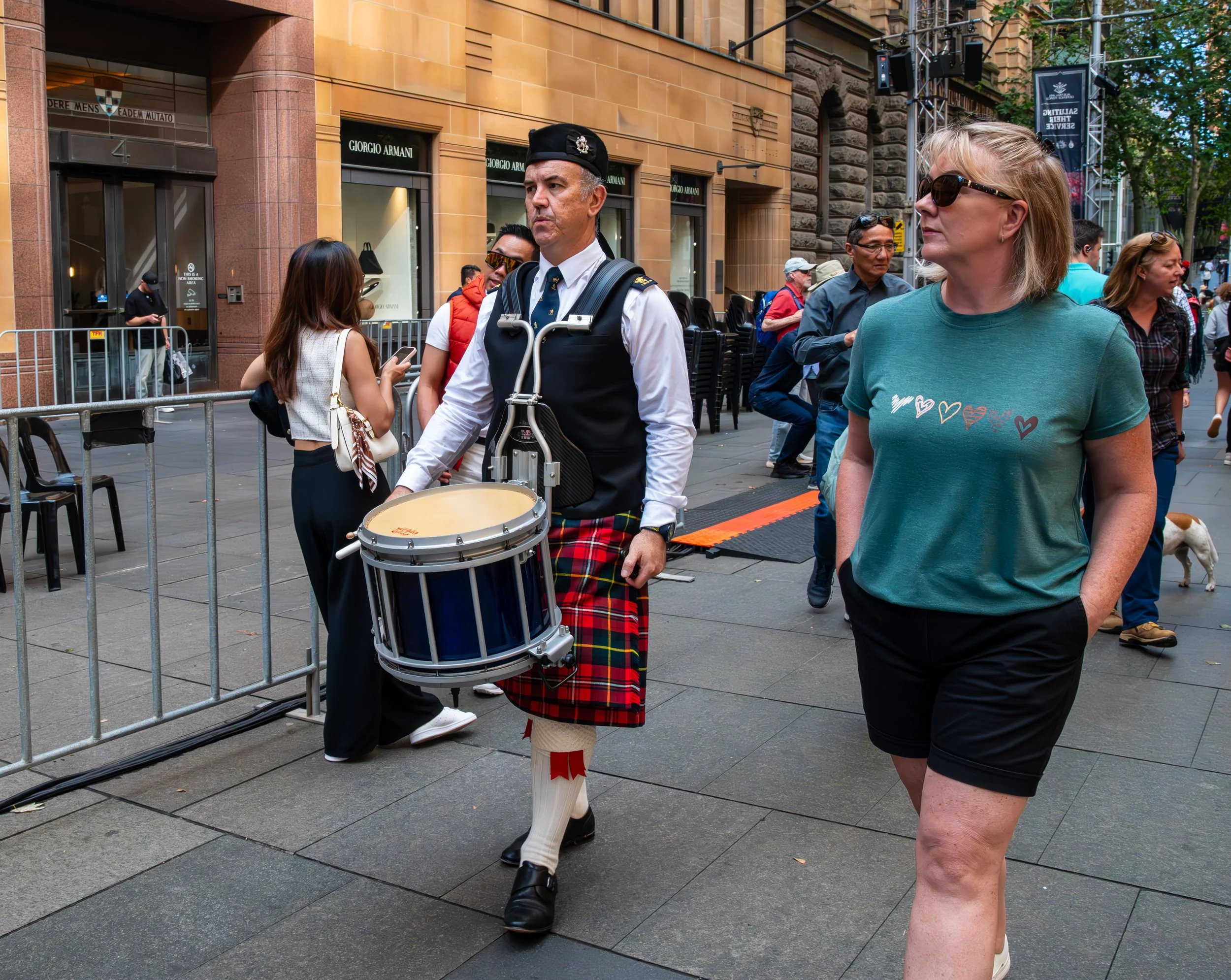 Street scene with a man in a kilt playing a snare drum and a woman walking nearby, others in the background, city buildings, and trees.