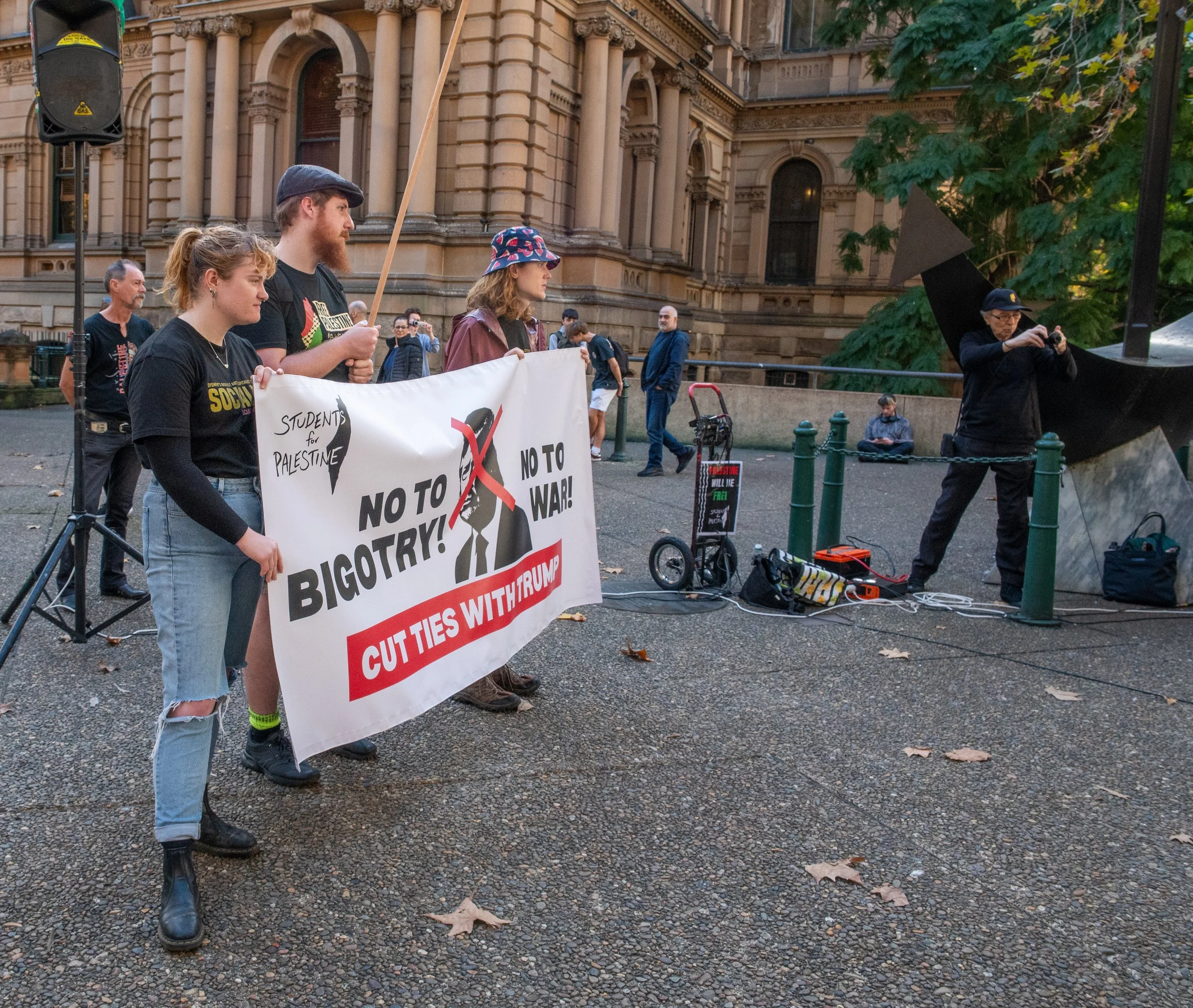 Group of protesters holding a large banner that reads 'No to Bigotry! No to War! Cut Ties With Trump' at a public demonstration. Several other people are visible in the background near a historic building, some sitting and some walking, with equipment set up on the street.