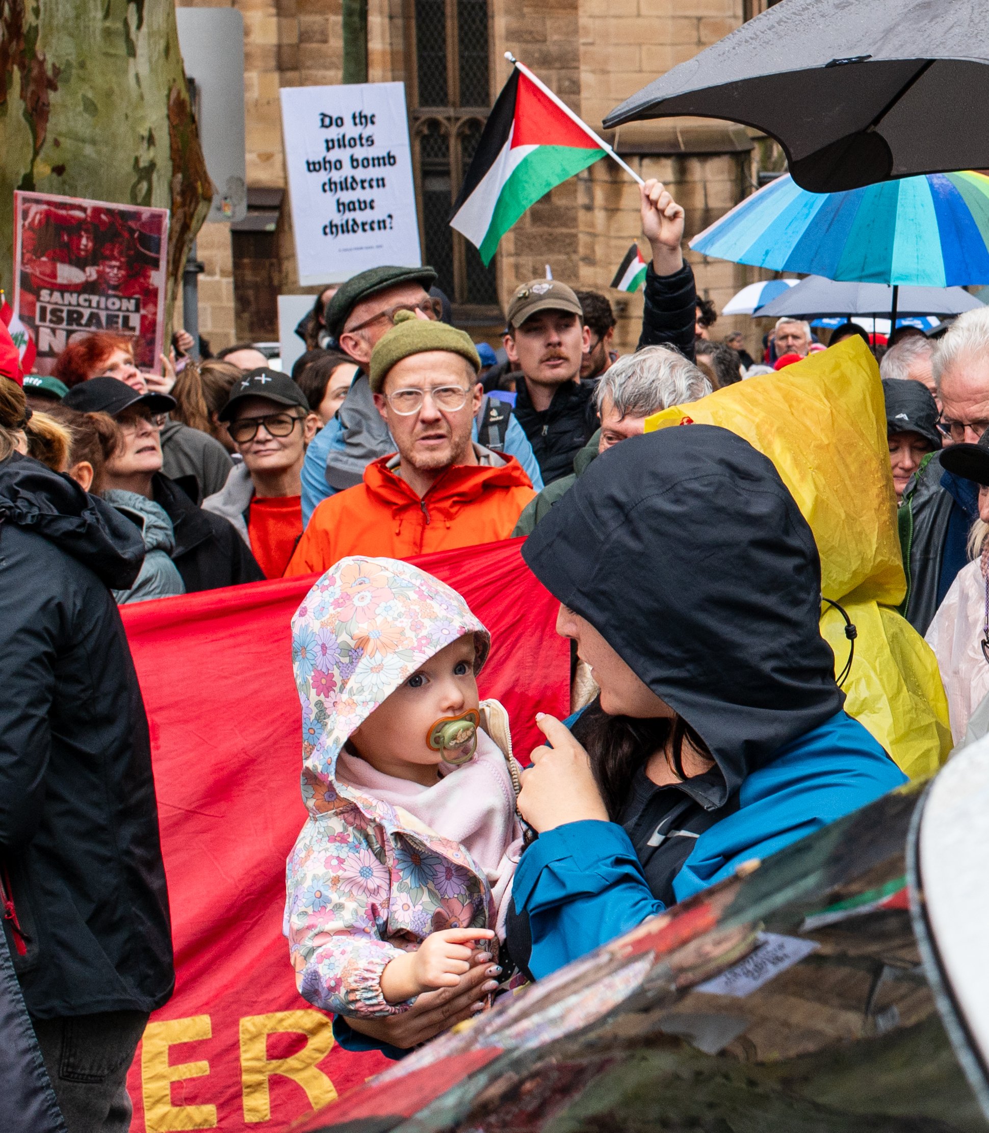 A crowd of protesters, some holding Palestinian flags and signs, gather outdoors in rainy weather, with many wearing rain jackets and holding umbrellas. One sign questions whether pilots who bomb children have children.