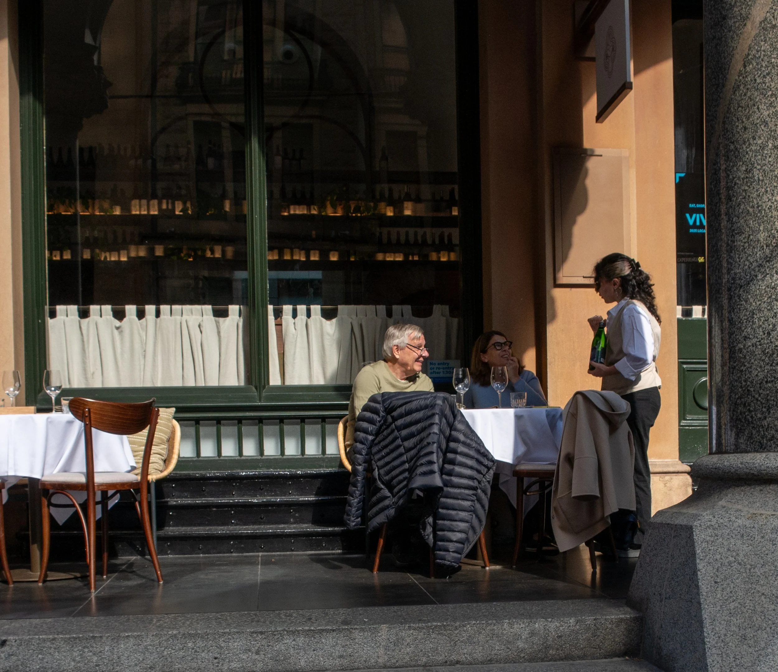 A woman serving drinks to two people dining outside a restaurant, with one person smiling and the other wearing glasses, set against a large window reflecting city buildings.