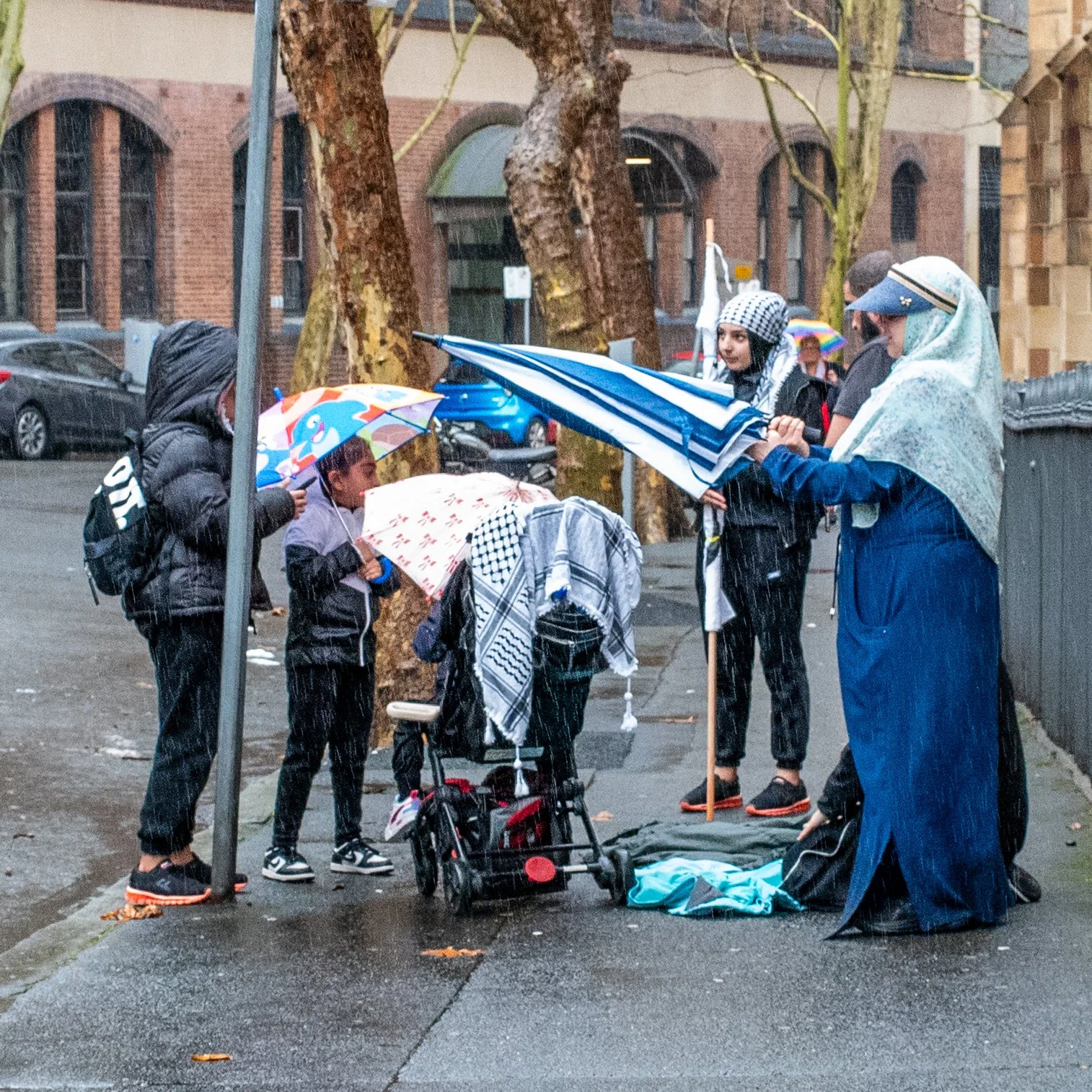 People standing under umbrellas on a rainy street, with a woman holding another umbrella and interacting with children and a person with a stroller, near trees and brick buildings.