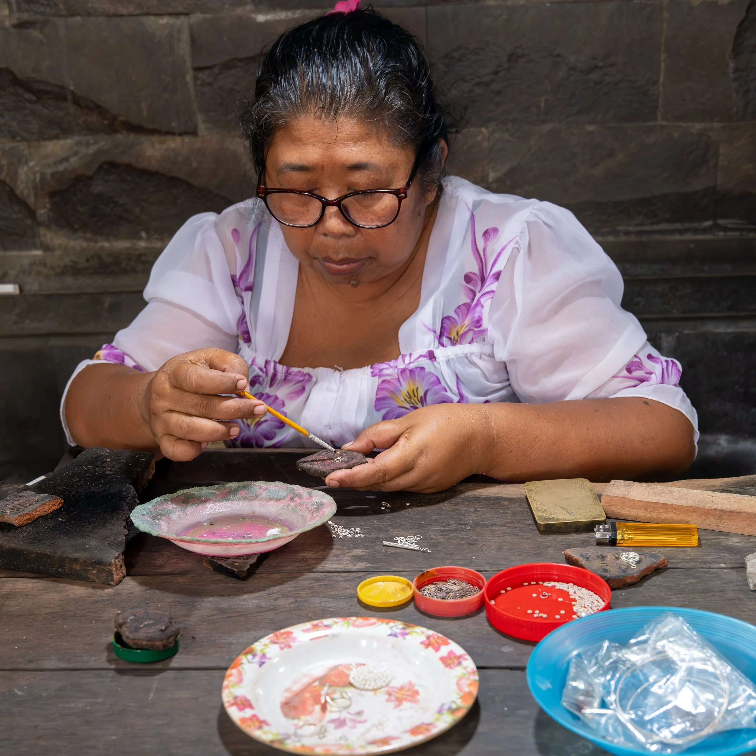 A woman with black hair, glasses, and a white dress with purple floral patterns is working on a craft project at a table filled with small containers of beads and jewelry supplies, outdoors against a stone wall background.