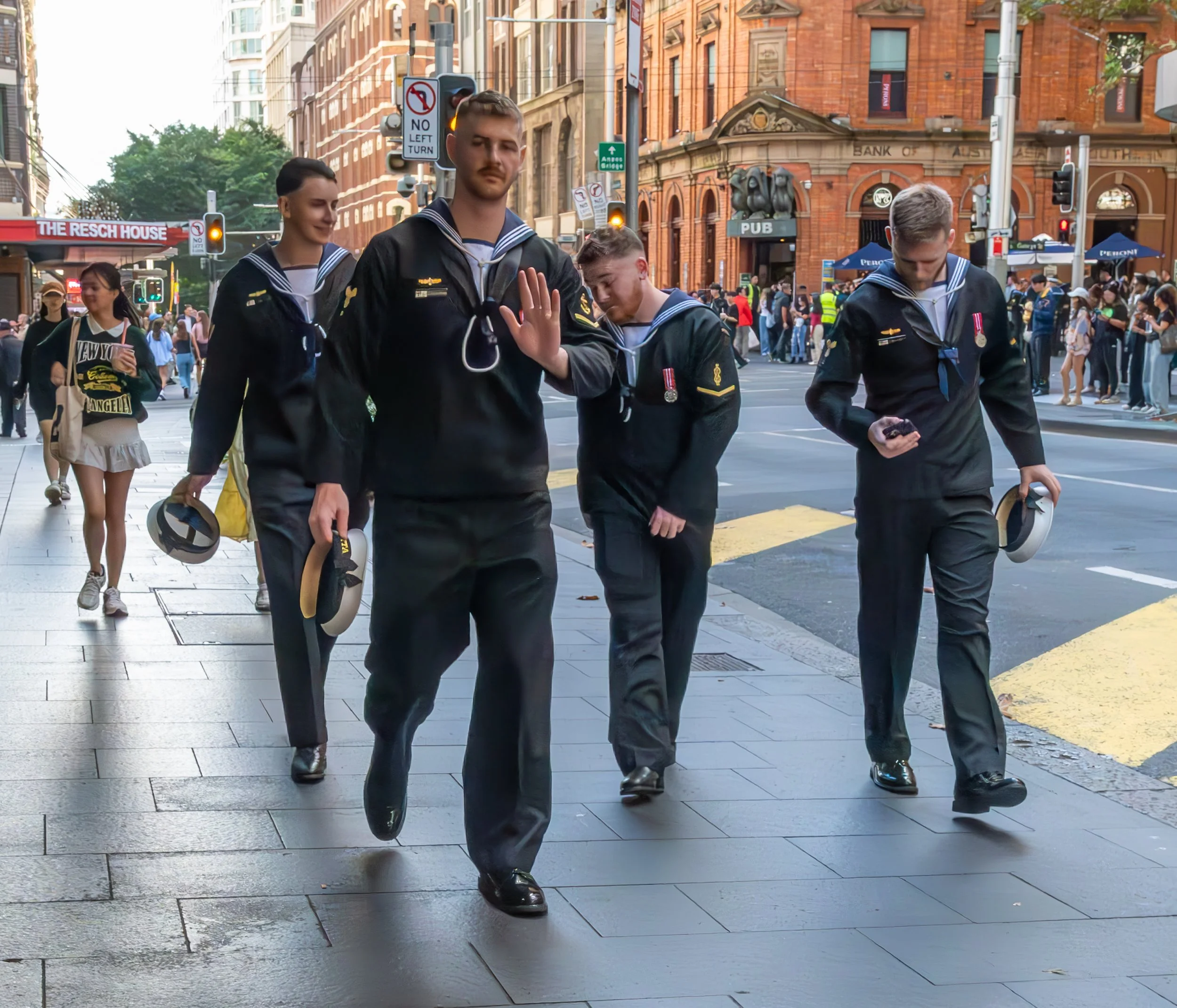 Four young men dressed in navy blue naval uniforms walk on a city sidewalk holding their hats. One of them raises his hand in a stop gesture. The background shows a busy street with pedestrians, a store, and historic buildings.