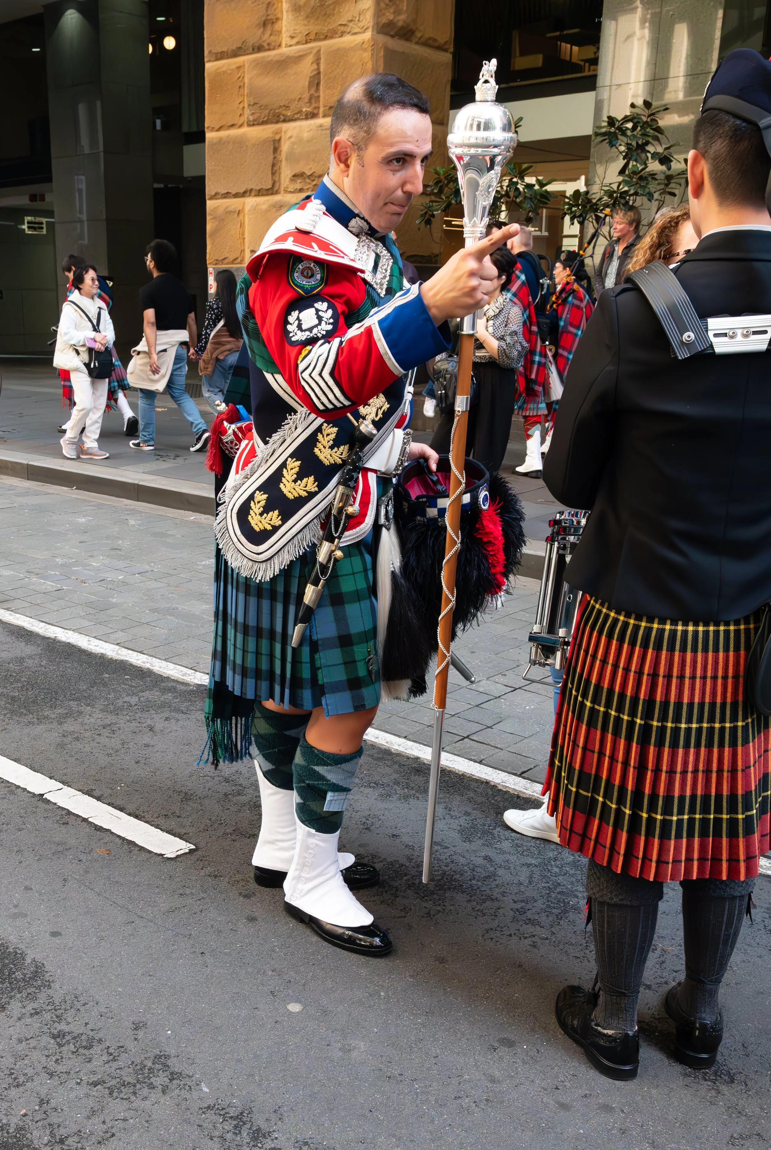 A man in traditional Scottish attire, including a kilt, sporran, and sash, standing on a city street and holding a ceremonial staff with a silver ornament on top, engaging in conversation with another person also dressed in a kilt.