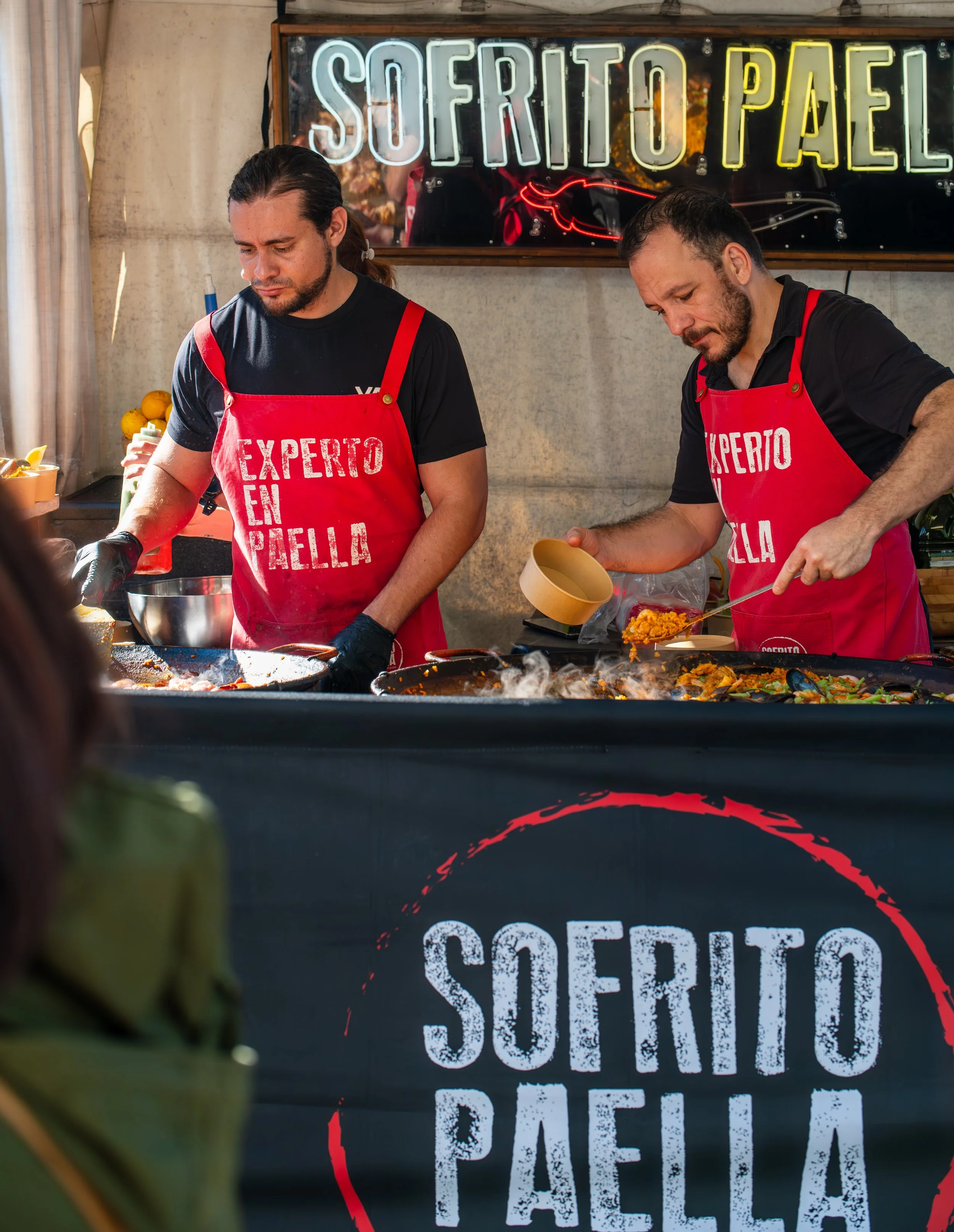 Two men cooking paella at a food stall, wearing red aprons that say 'Experto en Paella' with a neon sign behind them that reads 'Sofrito Paella'.