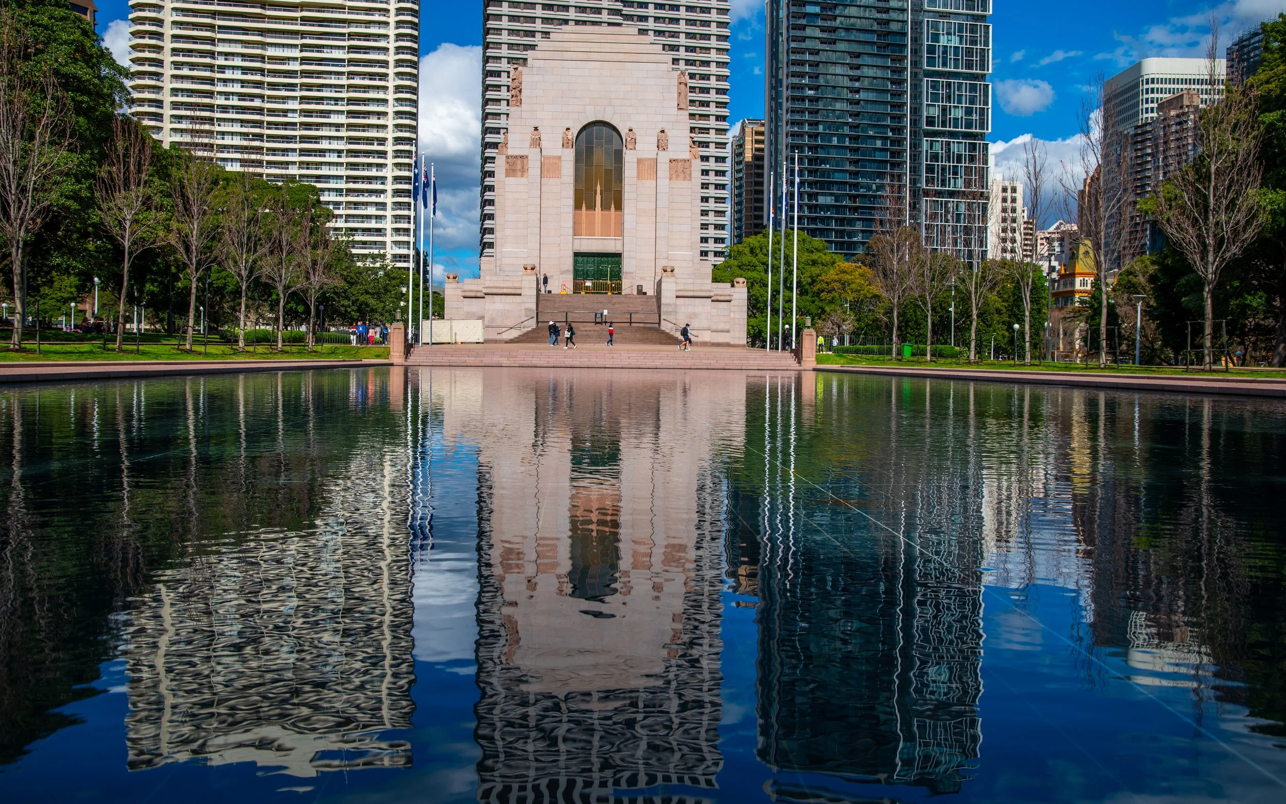 A city park with a reflecting pool in the foreground and a large stone monument at the center, surrounded by modern high-rise buildings and trees.