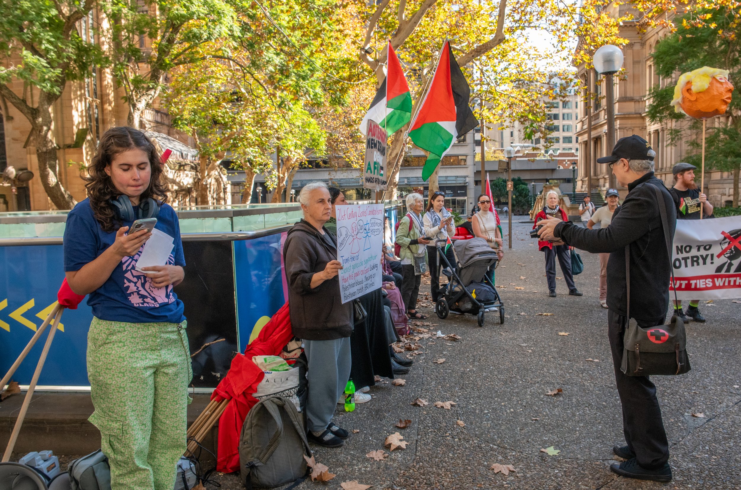 A group of people gathered outdoors on a city street during autumn, protesting with flags and signs. Several individuals are holding Palestinian flags, and one person is speaking into a microphone. The demonstrators include women and men, some with strollers, surrounded by trees with yellow leaves and city buildings.