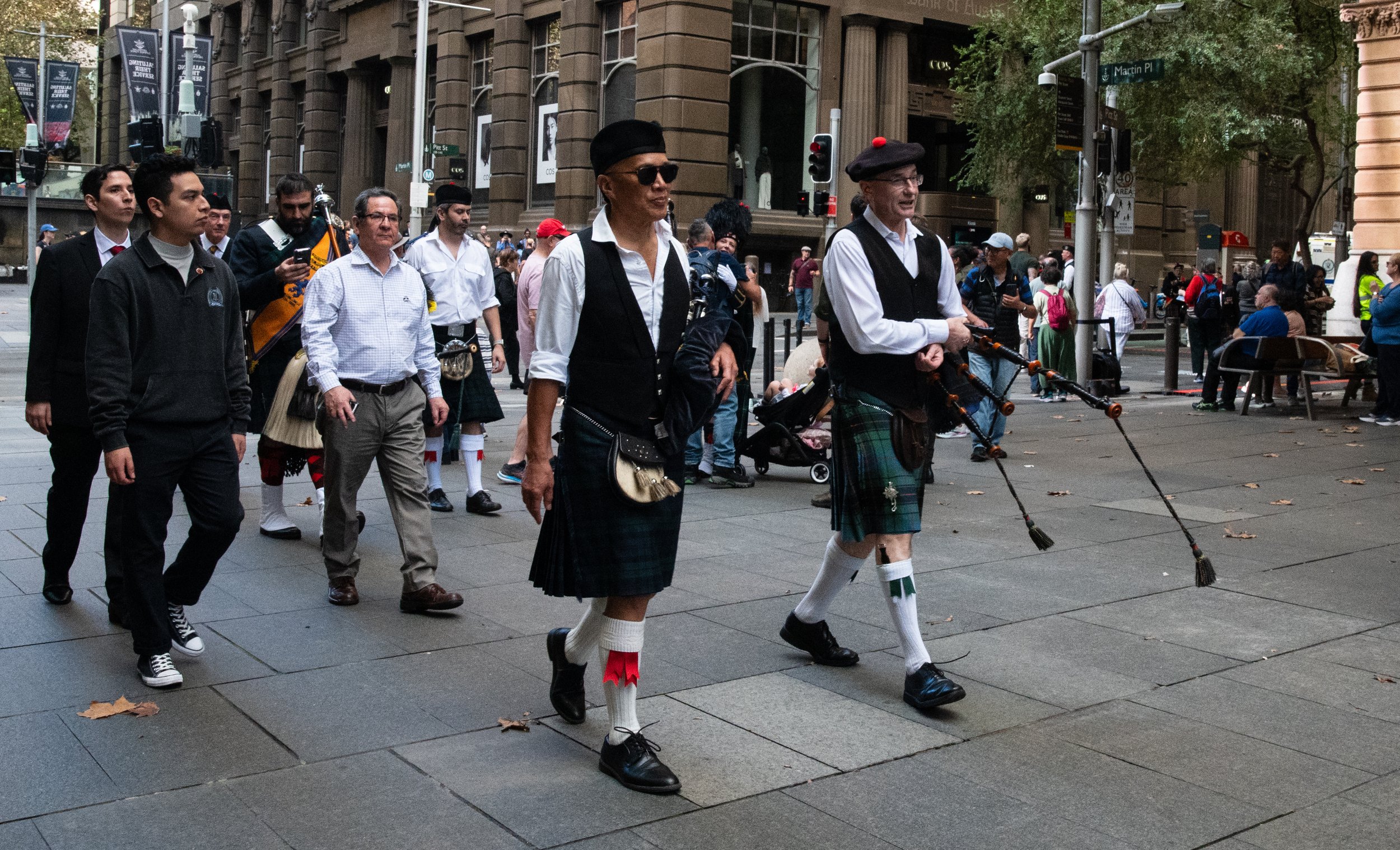 Group of people walking in a city crosswalk, some wearing traditional Scottish kilts and playing bagpipes, others in modern clothing, with tall buildings and trees in the background.