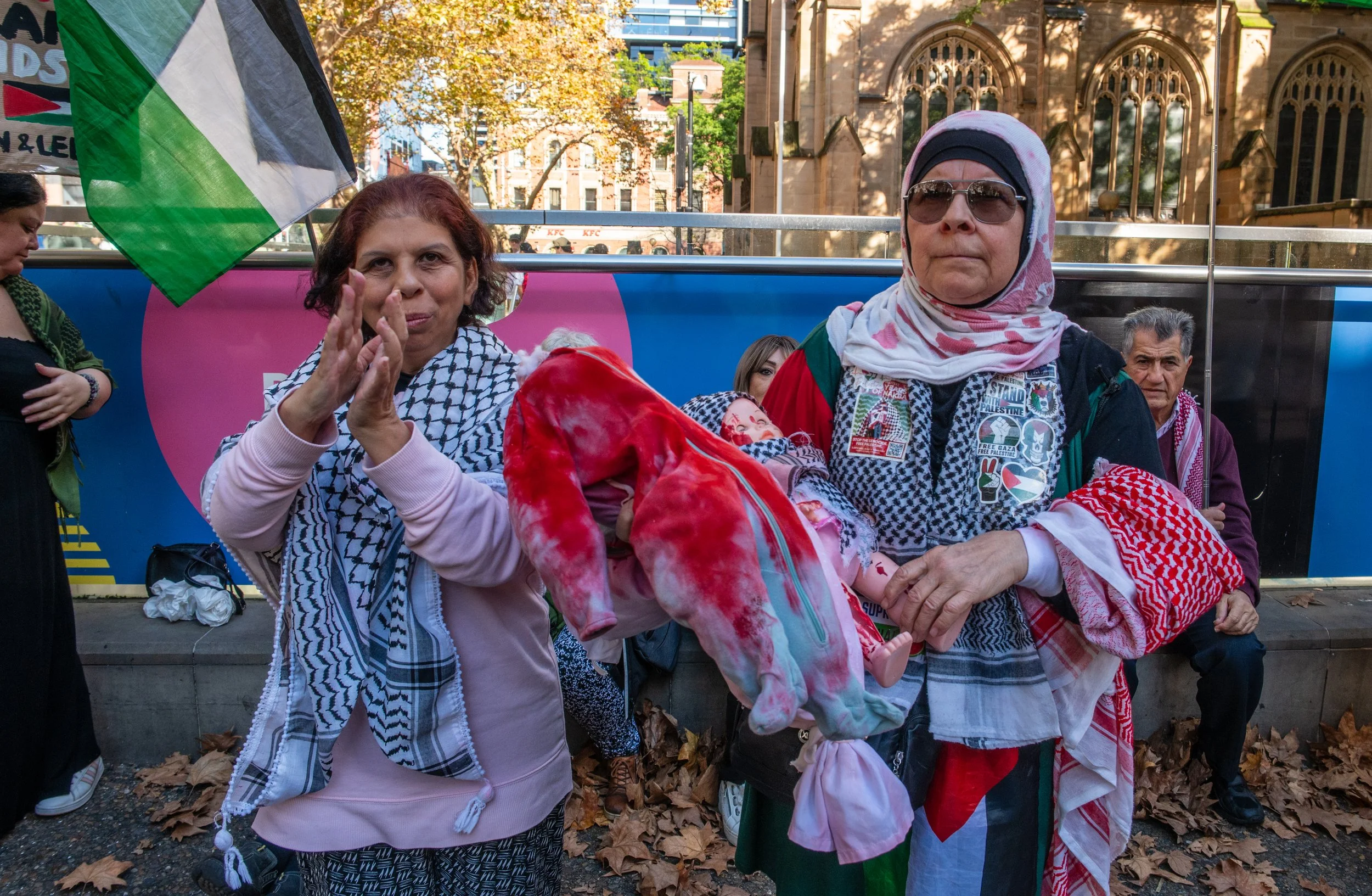 Two women hold a baby doll wrapped in a red and white cloth during a protest or demonstration, with people and a building in the background.