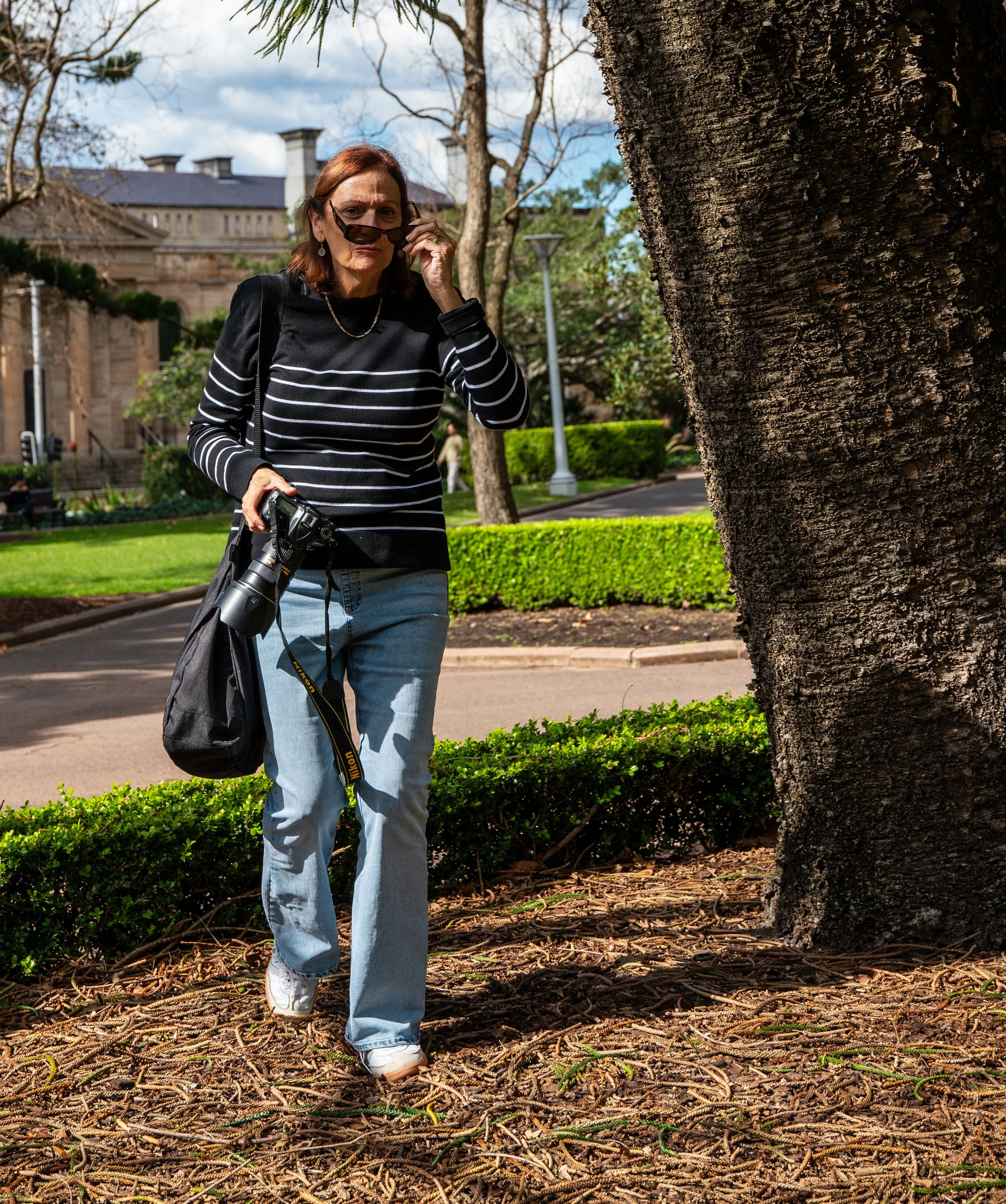 Woman with sunglasses and camera walking on a park sidewalk near a large tree with a building in the background.