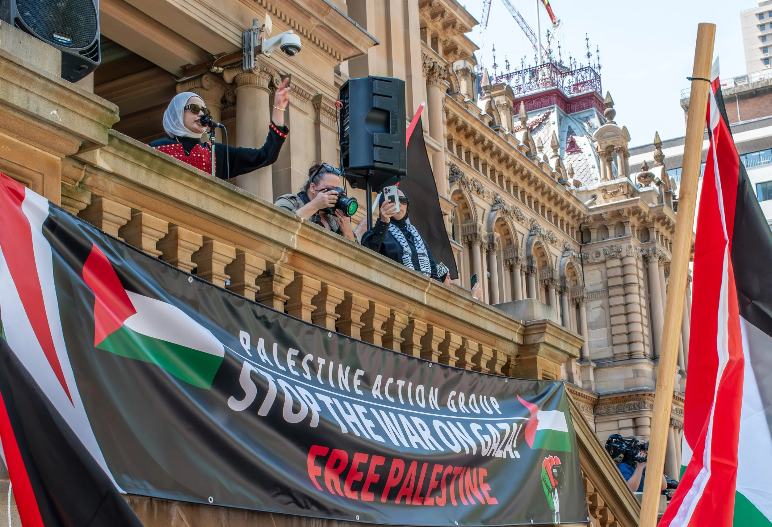 People on a balcony holding flags and banners, protesting for Palestine, with a large banner that reads 'Palestine Action Group Stop the War on Gaza, Free Palestine'.