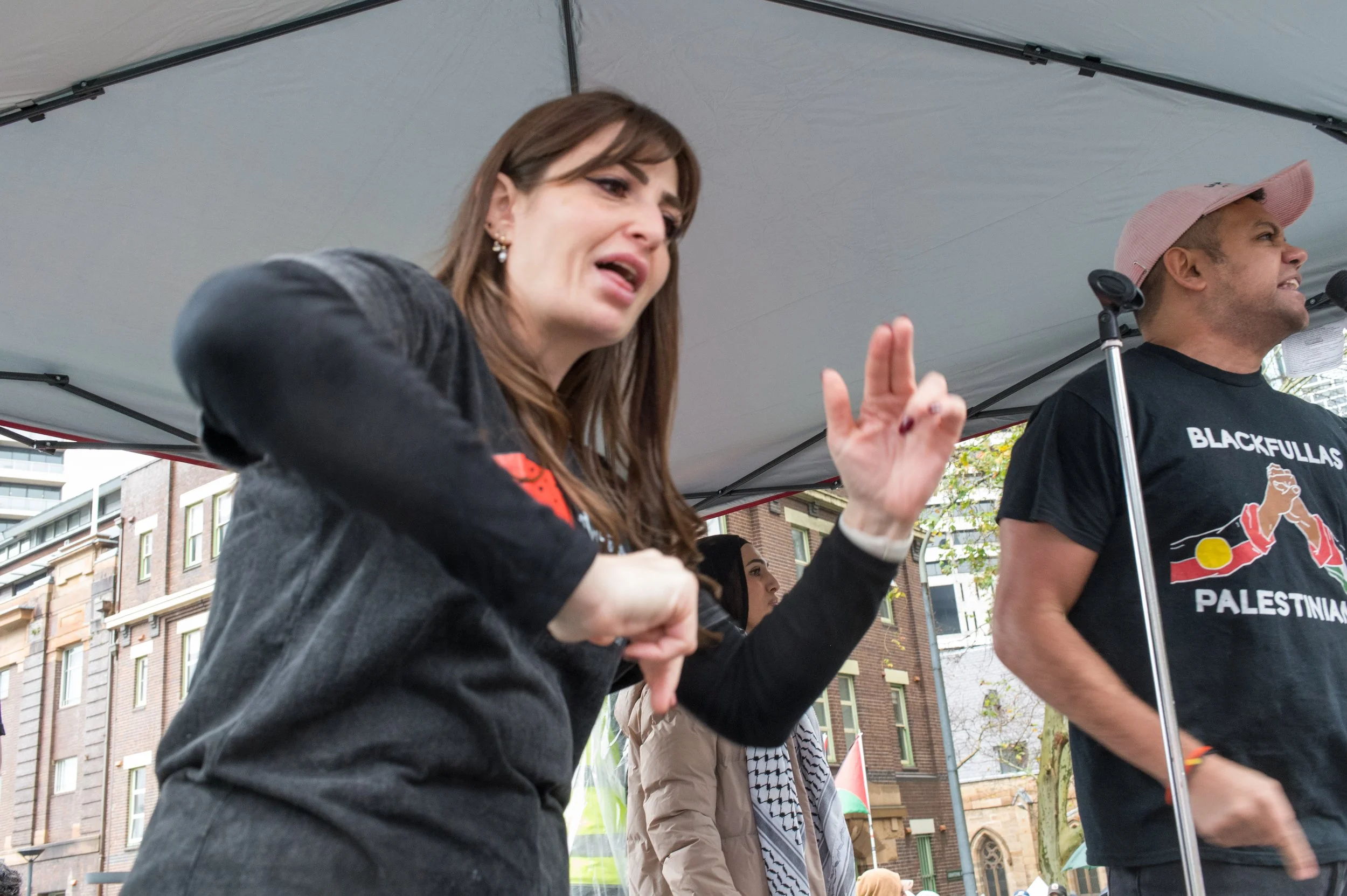 A woman speaking passionately at a rally, making a peace sign with her fingers. She is wearing a black jacket and has long brown hair. A man next to her is wearing a black t-shirt with the words 'Black Fullas Palestine' and a pink cap, standing at a microphone. Other people and buildings are in the background.