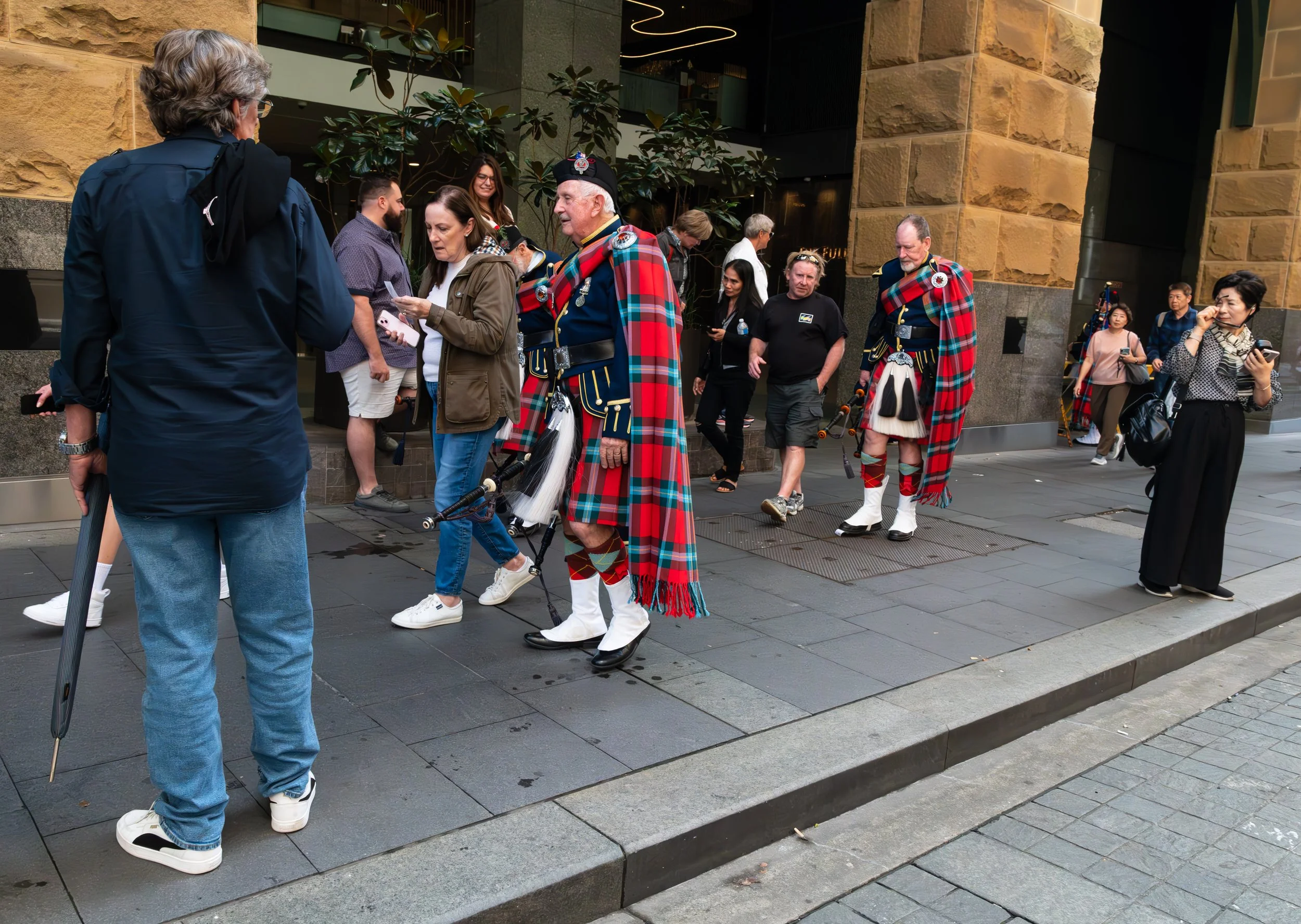 Tourists and bagpipers in traditional Scottish attire walking on a city sidewalk.