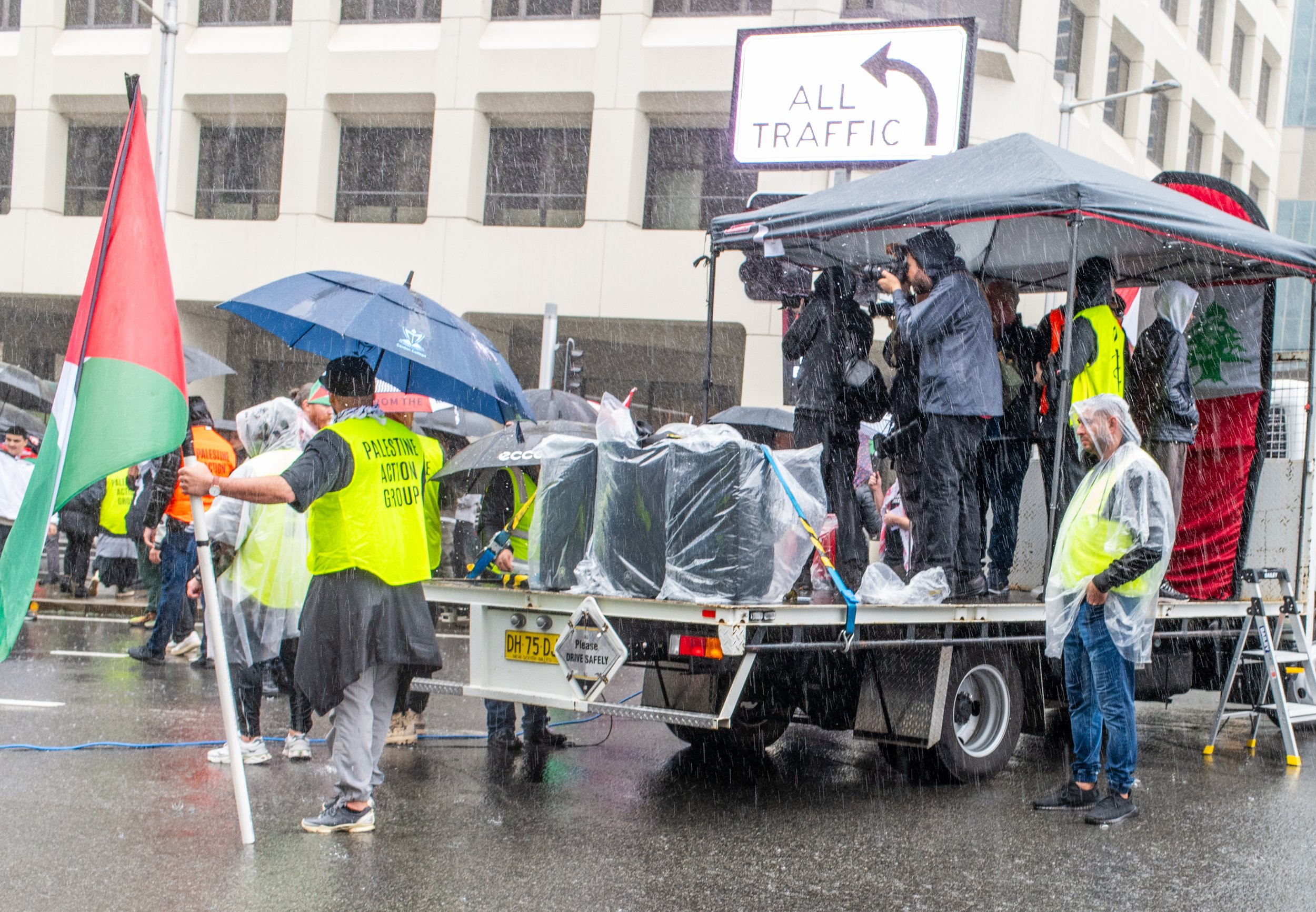 A group of people, some with yellow vests reading 'Palestine Action Group,' are participating in a protest in the rain. They are on and around a vehicle with a canopy, some holding umbrellas. One person holds a Palestinian flag, and some are filming or taking photos. The scene is set on a city street with a building in the background and a traffic sign indicating all traffic must turn left.