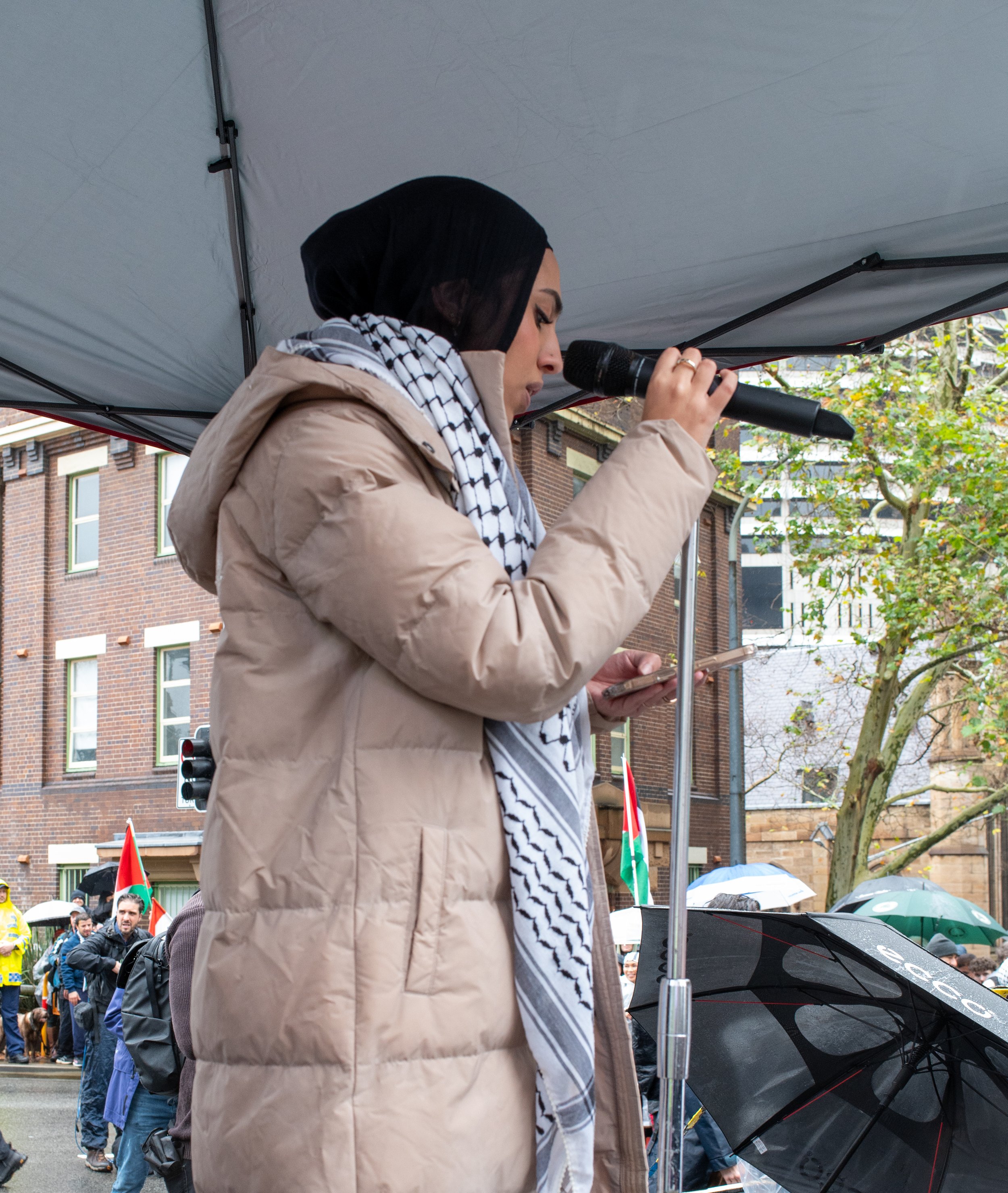 A woman with dark hair, wearing a beige coat and a black and white scarf, speaking into a microphone at an outdoor protest or rally, with people holding umbrellas and flags in the background.