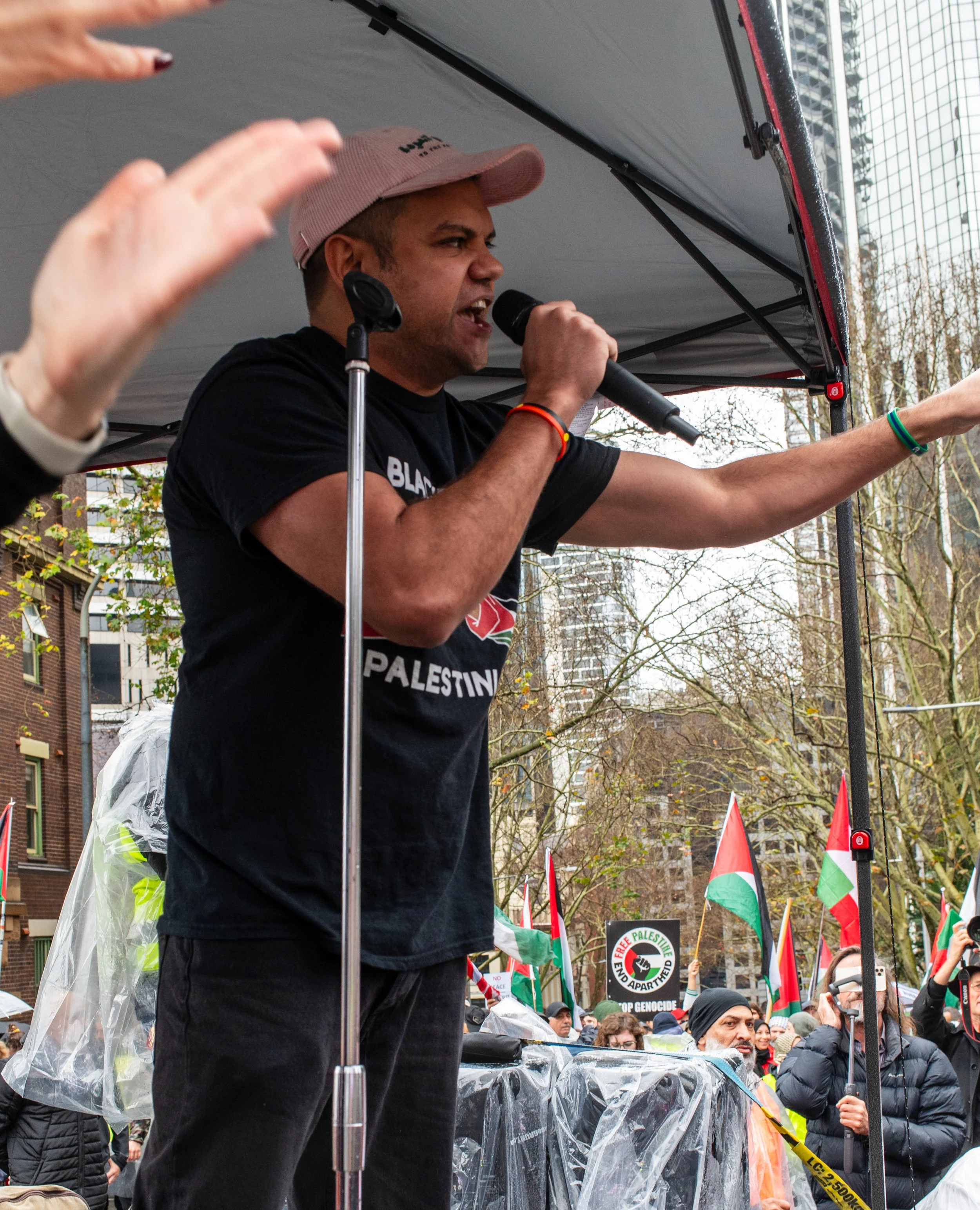 A person speaking passionately on a stage at a protest or rally, with a crowd holding flags supporting Palestine and signs, in an urban outdoor setting with tall buildings and leafless trees.