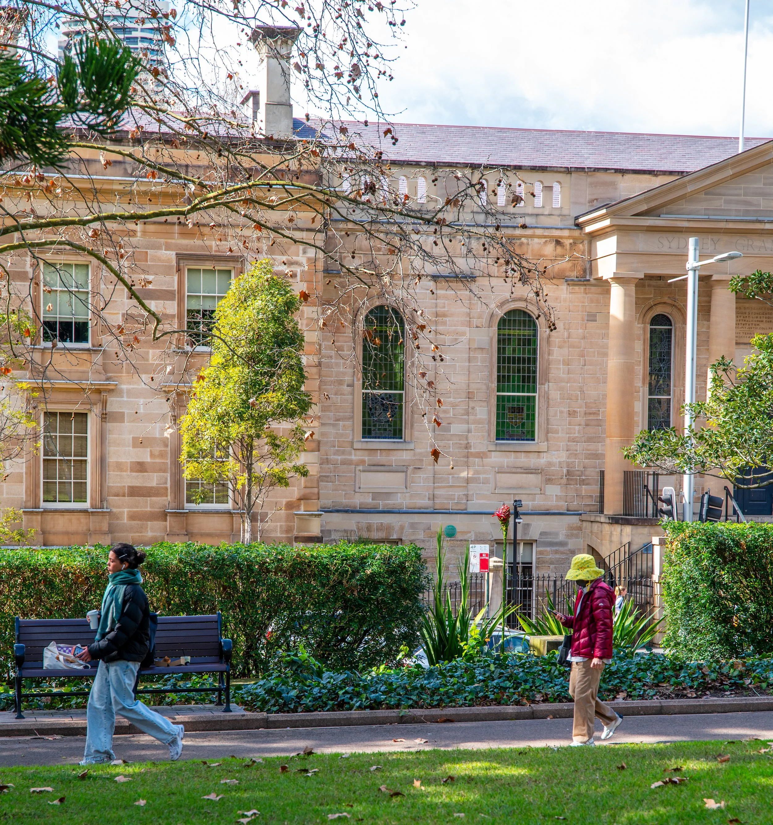 Two people walking in a park on a sidewalk, with a historic sandstone building with arched windows and stained glass in the background. One person is wearing a black jacket and light blue jeans, holding a takeout container. The other person is wearing a yellow hat, red jacket, and beige pants, looking at their phone. There are trees, green shrubs, and a bench along the sidewalk.